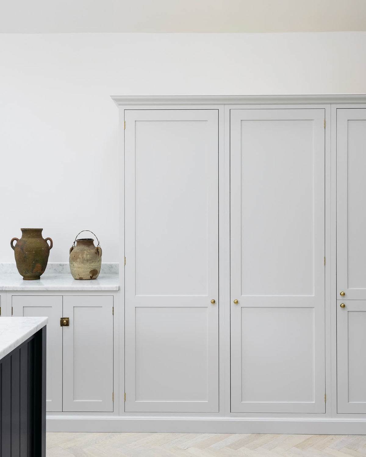 A spacious and artisan Shaker kitchen features an off-white L-shaped sink run with a Carrara marble worktop, a large pantry-blue kitchen island, and a bank of full-height cupboards. Cleverly decorated with vintage ceramics serving as artistic displays, it creates a simple yet intriguing kitchen space.