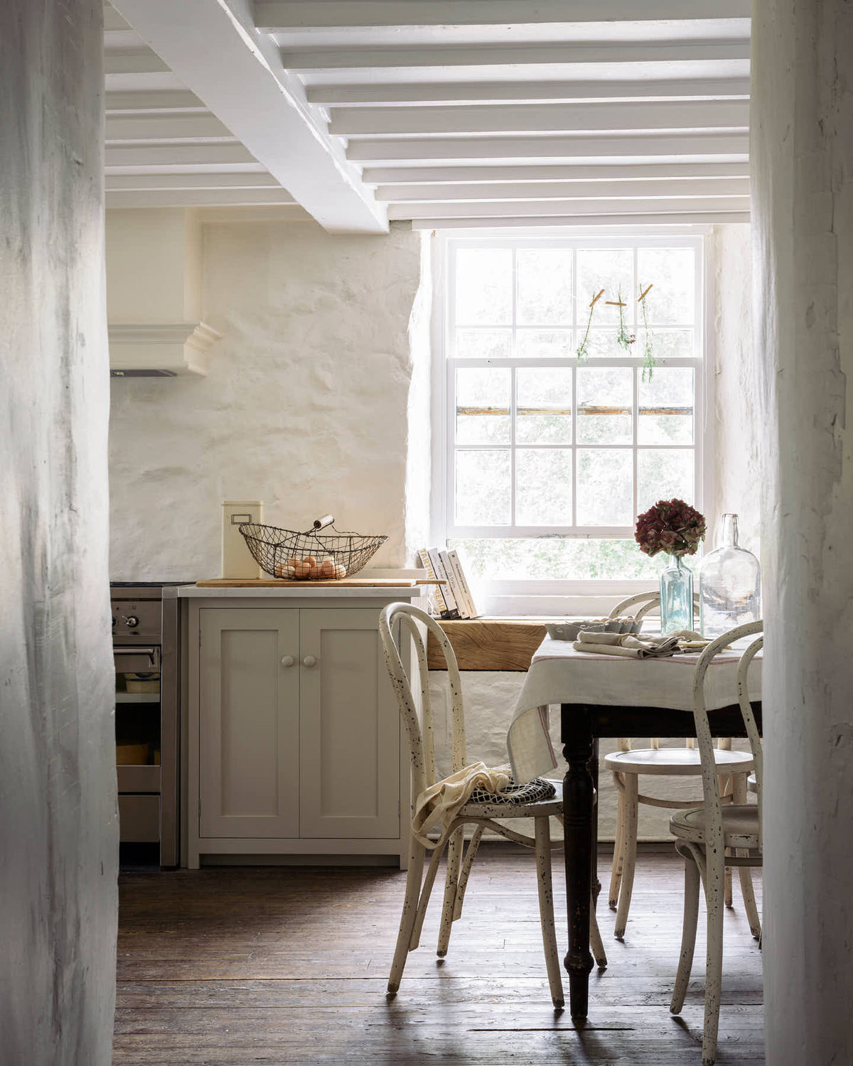 An atmospheric image of bright white light filling a simple English farmhouse-style kitchen with assorted vintage furniture, located at deVOL’s headquarters in Cotes Mill. The room features low, exposed wooden beams and worn wooden floorboards.