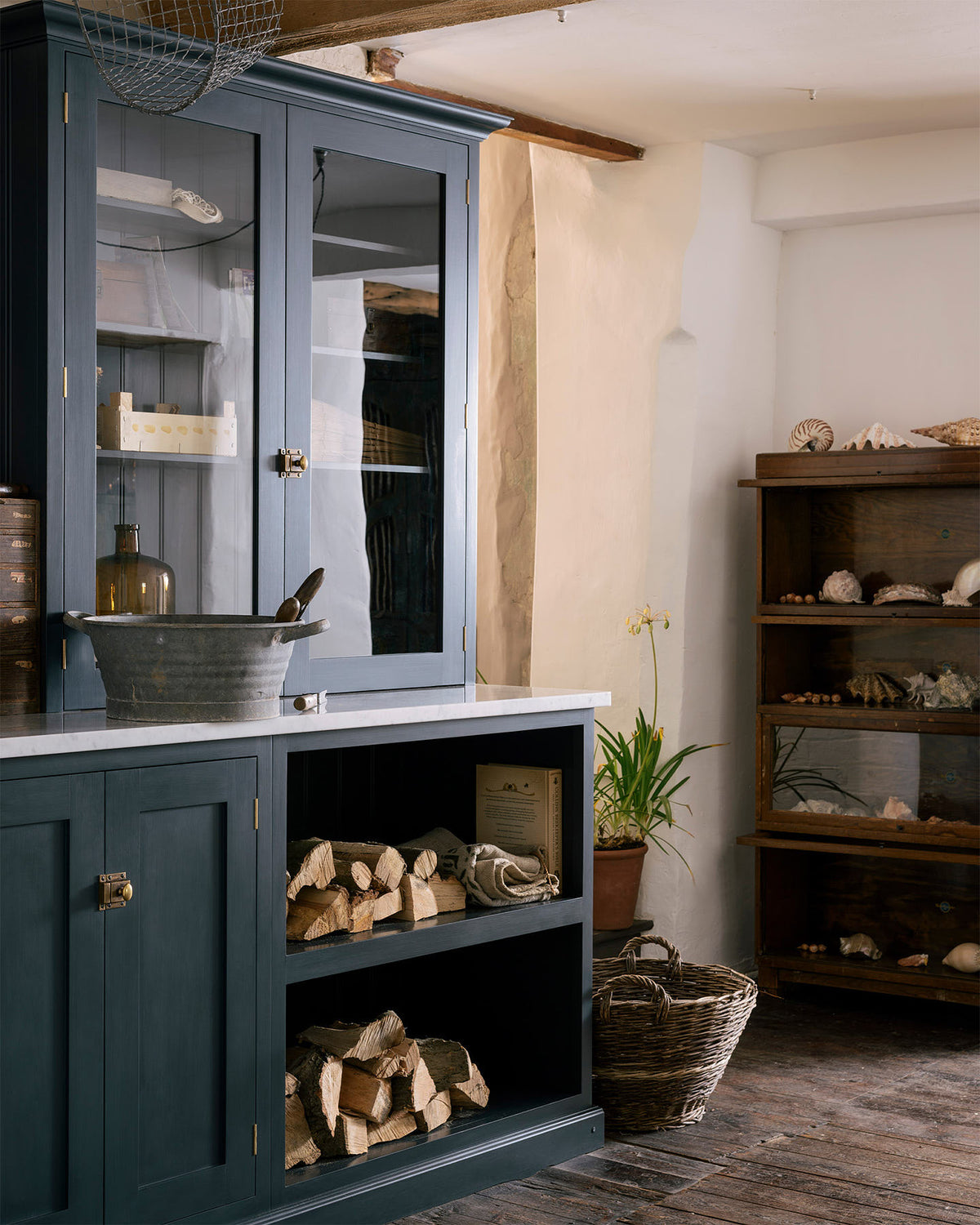 An angled look at the wooden and metal accents—including firewood, a metal bin, and a wooden toolbox of the pantry blue shaker kitchen’s sink run features a shelf, countertop glazed cupboards, a mix of closed and open base cabinets and a Carrara marble worktop.
