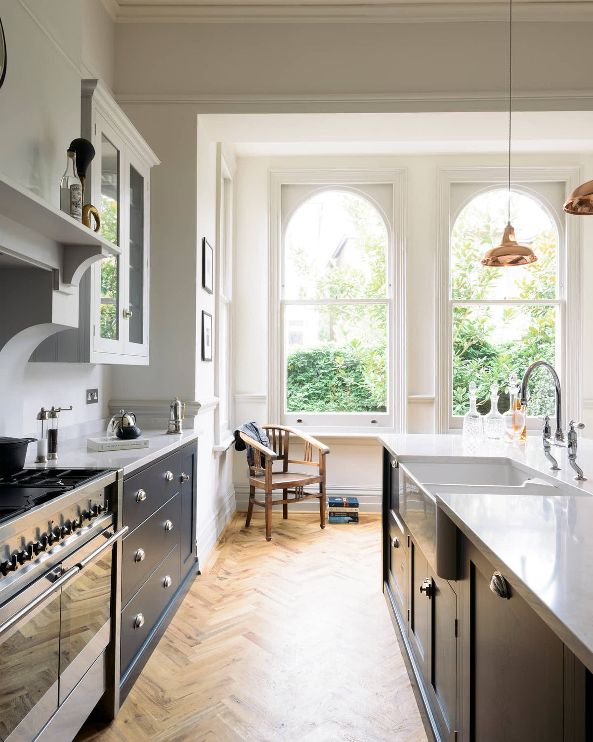 The pantry blue shaker kitchen features creamy Silestone worktops. Bright sunshine streams through the tall arched window, illuminating the central island, which houses a sink on one side and a breakfast bar on the other.
