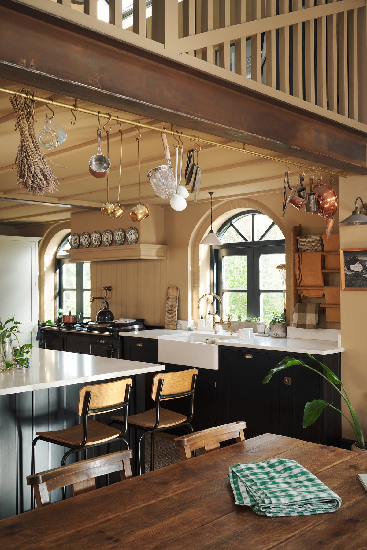 An oblique view of a large, open Printer’s Black Shaker kitchen. A long cooker and sink run is topped with a Silestone worktop, complemented by both an open and closed pantry larder, and a kitchen island. Decorated with a show-stopping heirloom gaselier light, antique furniture, and an aged brass pot filler, the room feels carefully considered, comfortable and highly functional.