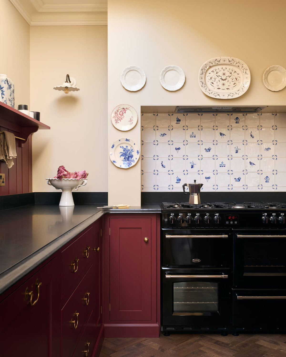A straight-on shot of kitchen, shaker-style cupboards are painted in deep red and run down the length of the left-hand side of the photo, they form an L and flank a black range cooker. The cooker splashback is made up of white and blue Delft tiles, and there are vintage-looking decorative plates around the chimney breast. The worktop is a black granite and has a decorative edge. The walls are a soft cream colour and the floor is dark brown parquet.
