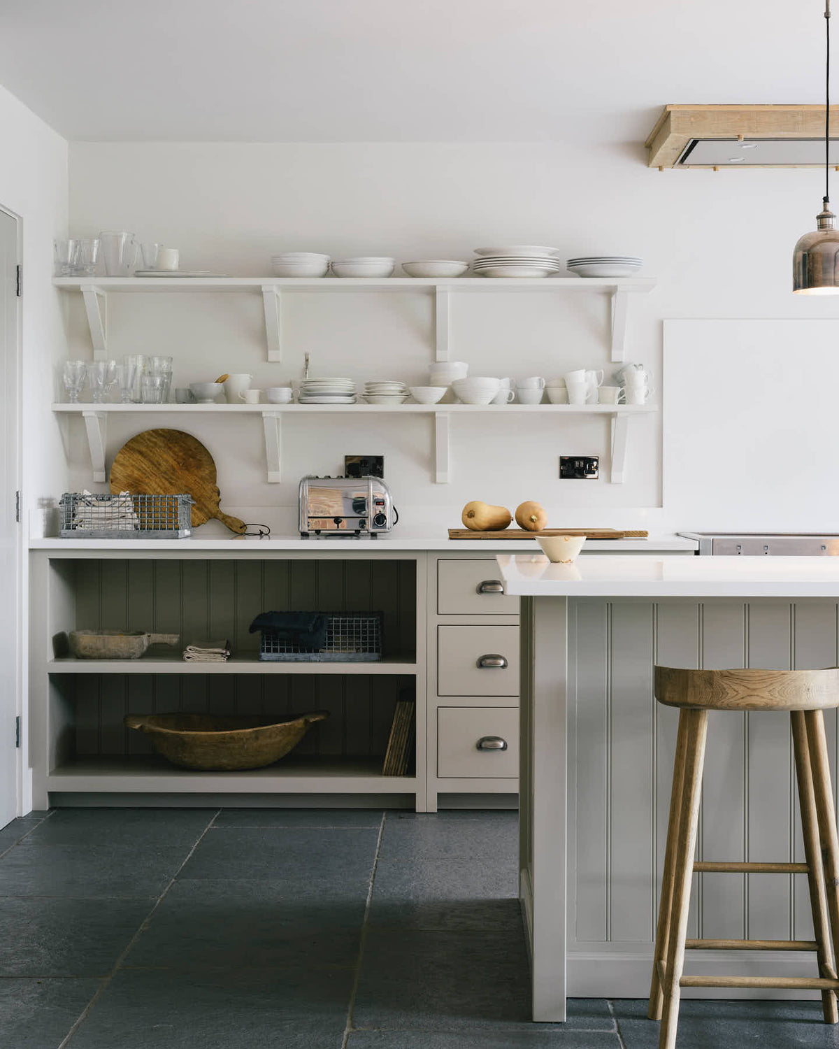 A quiet corner of a tranquil and calm Shaker kitchen features a long sink and cooker run with a Silestone worktop and splashback, a long kitchen island with a breakfast nook, and shelves. Beautifully decorated with a vintage stool that matches the raw wooden beams, muted ceramics, and silver pendant lights, this kitchen creates a lovely, simple space that embraces natural materials.