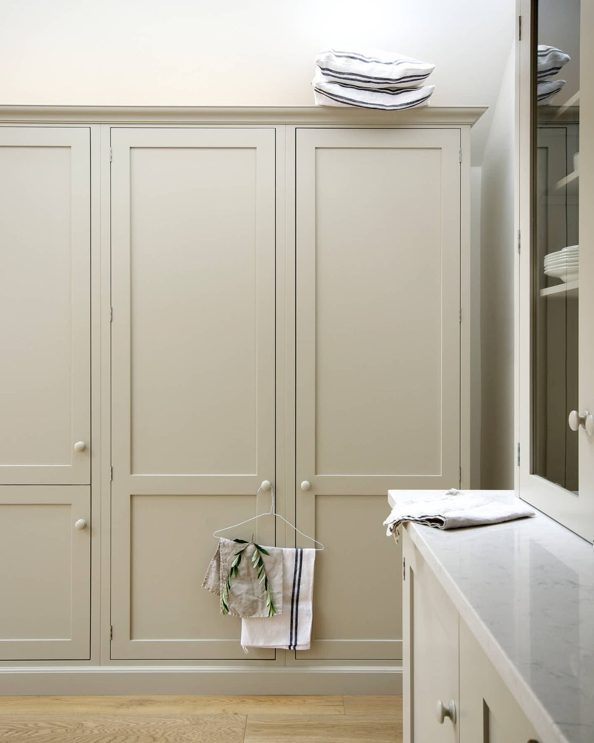 A corner of an elegant mushroom-coloured Shaker kitchen features a sink and cooker run with glazed countertop cupboards, and a bank of full-height cupboards. Adorned with simple plates, tea towels, pillows and pillow cases, it creates a calm and functional space.