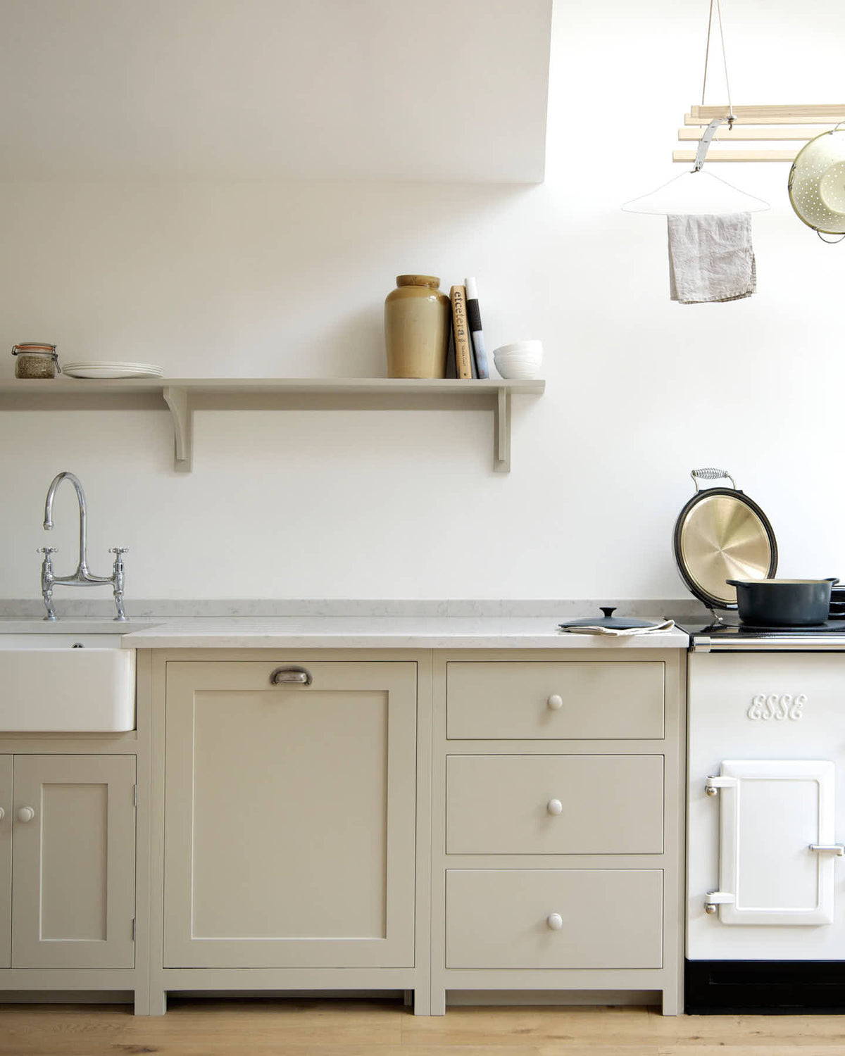 An elegant mushroom-coloured Shaker kitchen features a sink and cooker run with a Carrara marble worktop, a white gas cooker, and bifold patio doors leading to the luscious garden. Adorned with cast iron pots, artisan ceramics, and a deVOL laundry maid, it creates a calm and functional space.