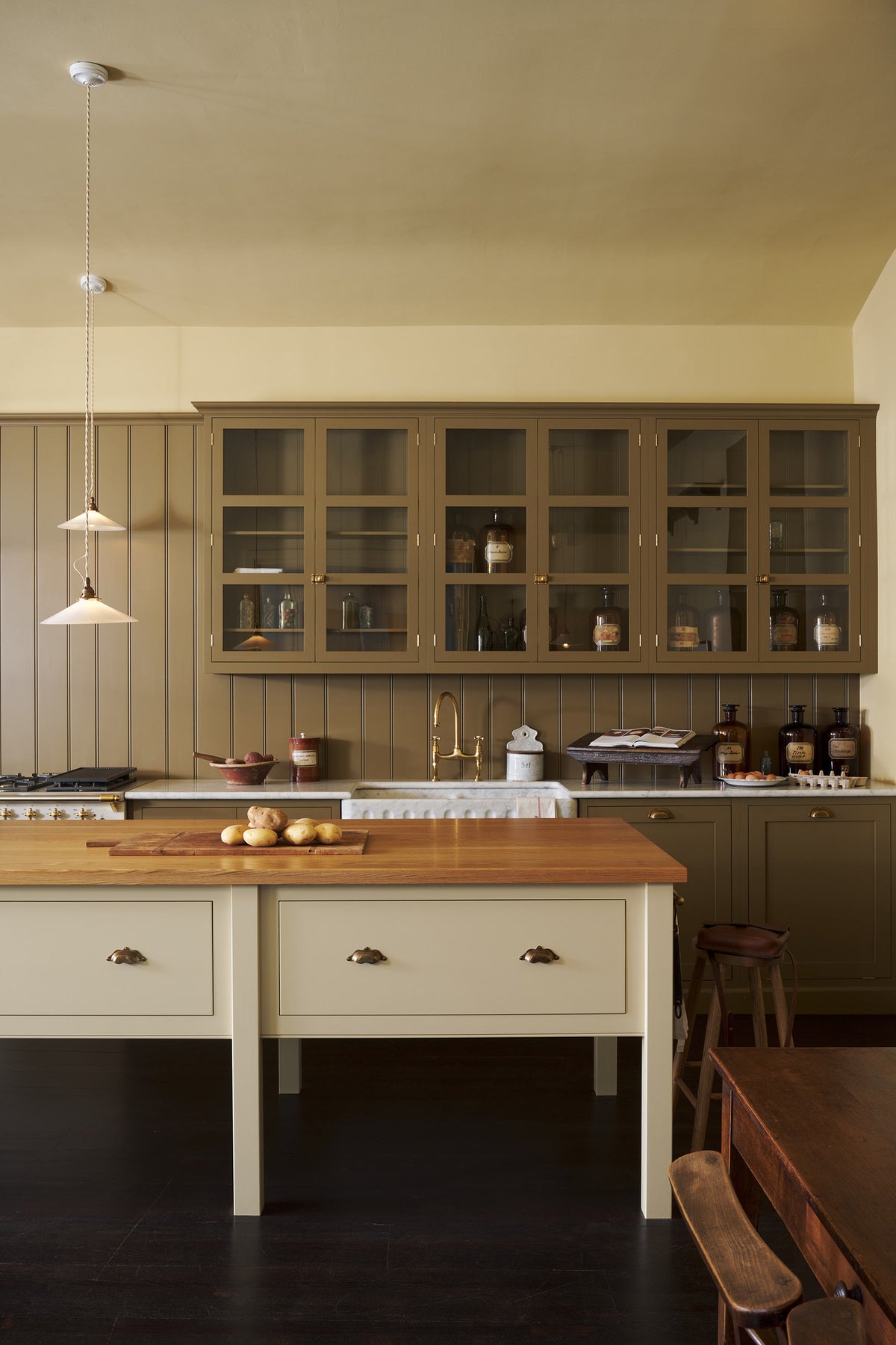 A large yet humble shaker kitchen features a long sink and cooker run with a marble worktop, a prep table with an oak worktop, a double fluted marble sink, glazed wall cupboards and a variety of antique furniture. The warm creams and greens makes the kitchen feel warm and inviting.