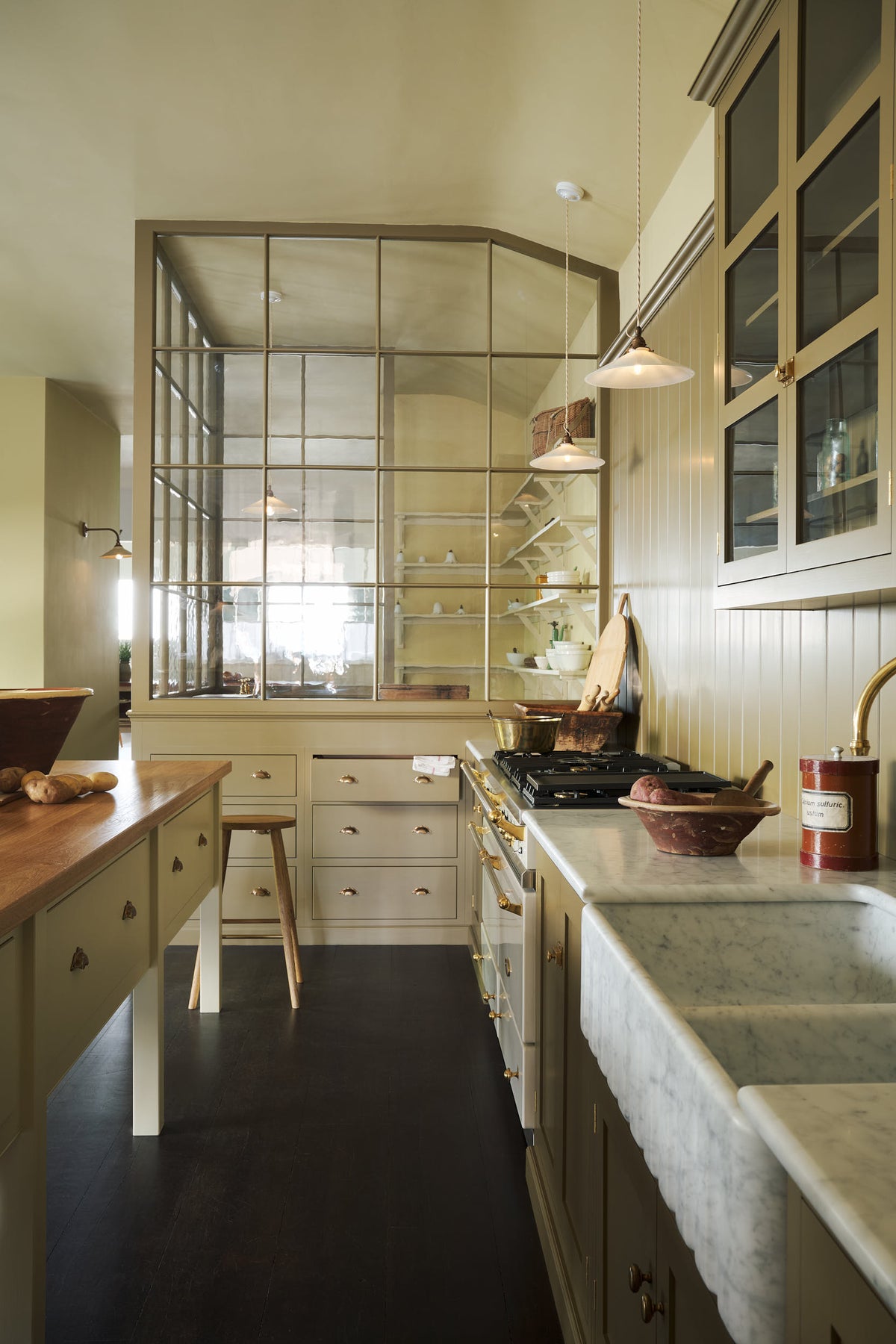 A closer look at a simple Quaker cream and Folgate green Shaker kitchen features a cooker and sink run with Carrara marble worktop, pan drawers, and a bespoke prep table with a classic oak worktop. Lovingly adorned with a tea towel, potatoes, antiques, and a Helen stool, it creates a warm and inviting atmosphere.