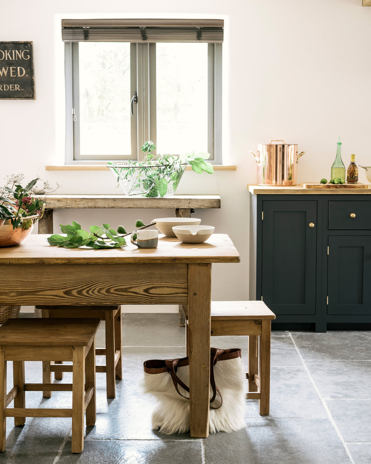 A closer look at the charming Pantry Blue cottage Shaker kitchen reveals a cooker run, open base cabinets, and a solid oak worktop that complements the wooden ceiling beams and vintage dining table with stools. The open cabinets display rose-gold cookware, reflecting the warmth of the wood while providing easy, functional access.