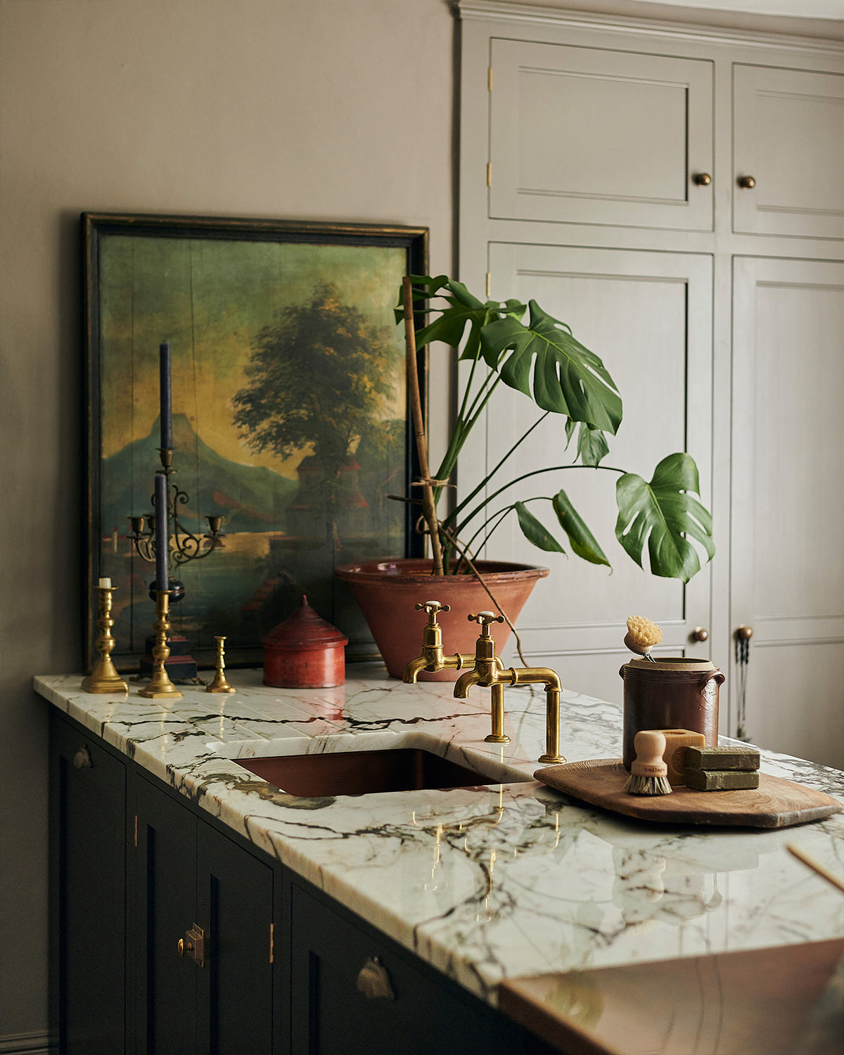 A closer look at a painter’s black shaker kitchen’s sink run with an aged copper sink, aged brass taps and a San Simone quartzite worktop - this stone has a pale white base with bold dark grey veining and little flickers of gold throughout. The walls are painted grey, and there is a cheese plant and a vintage oil painting in the background.