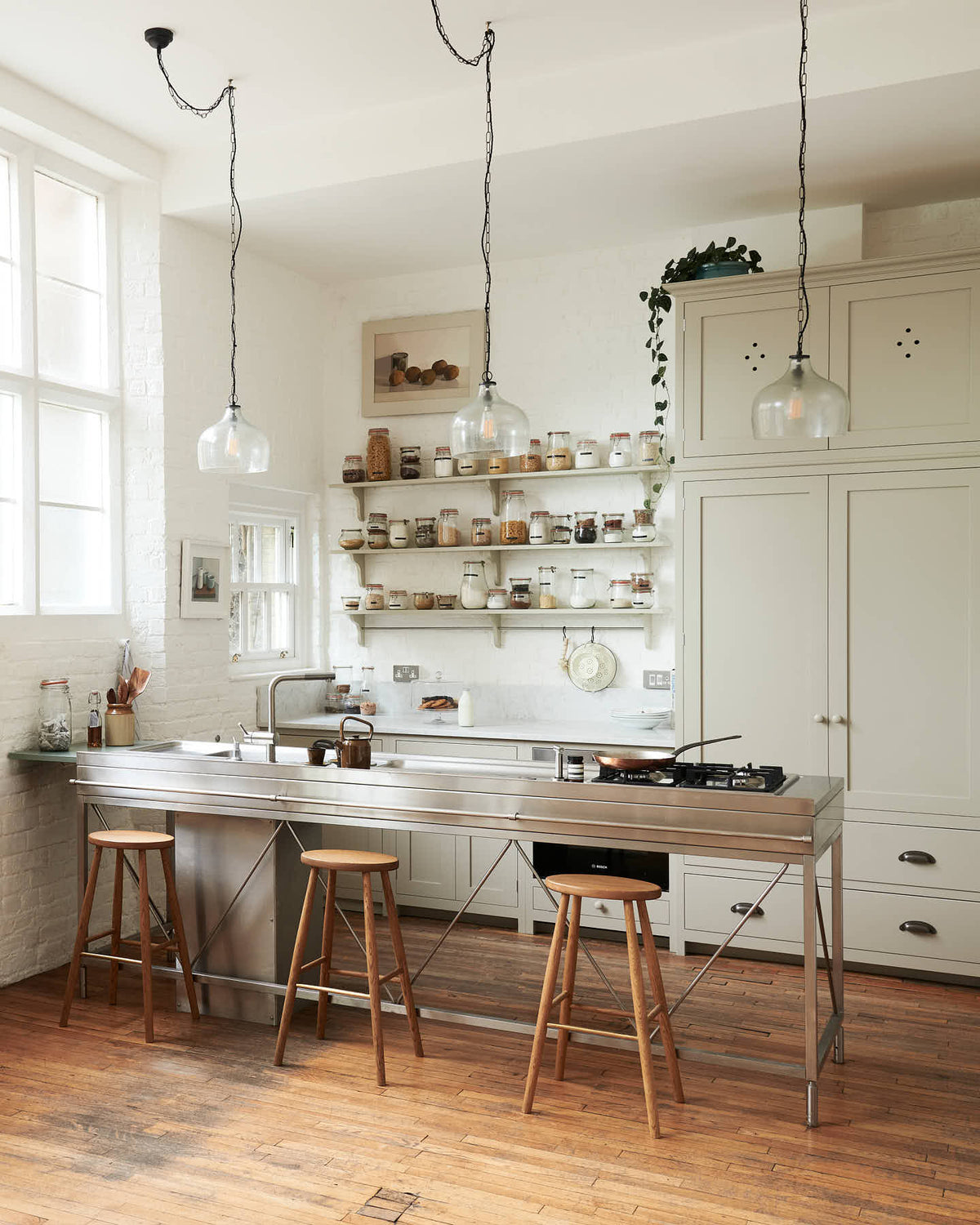 An oblique view of a tall and simple mushroom-coloured kitchen features a Shaker cupboard run with a full-height cabinet, a large steel cooker and sink kitchen island, and shelves with hanging rails. Adorned with Helen stools, glass jars of dried ingredients, an amber glaze teapot, glass and steel pendant lights, a silver fridge, and still-life paintings, it creates a humble industrial kitchen space.