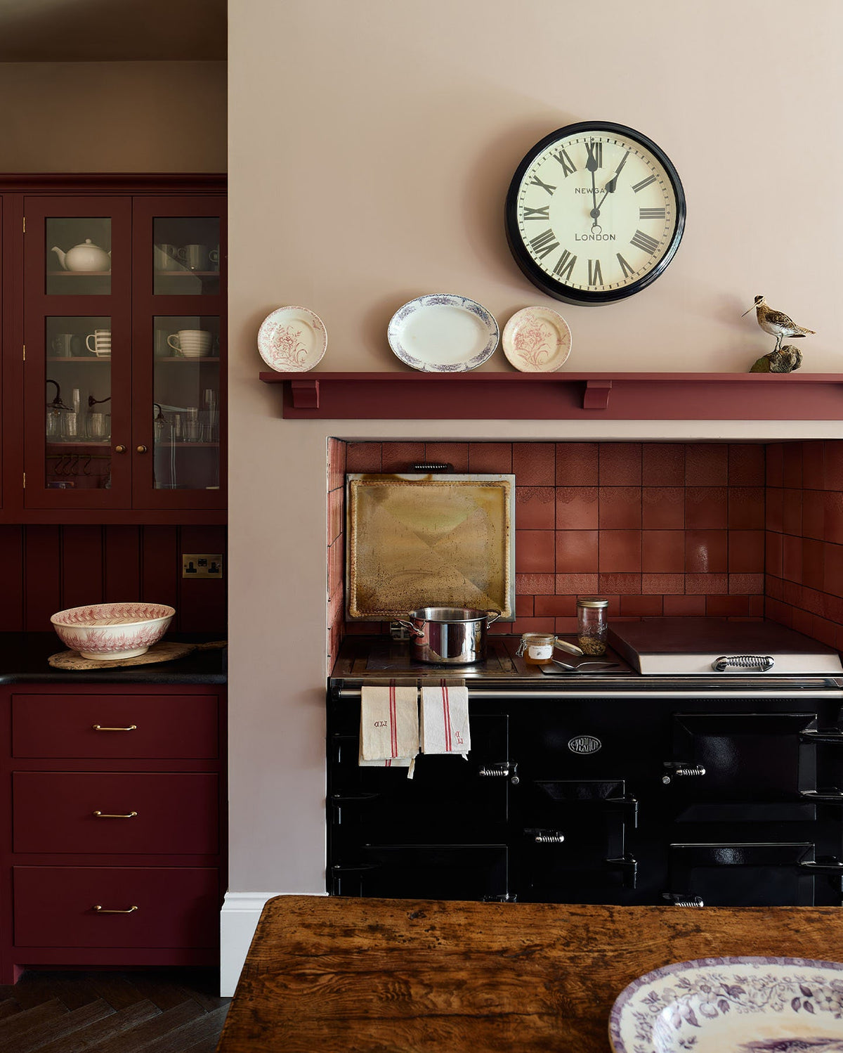 A straight-on view of a red painted Shaker-style kitchen with a large black Aga cooker in a chimney breast alcove with glossy red patterned wall tiles above.