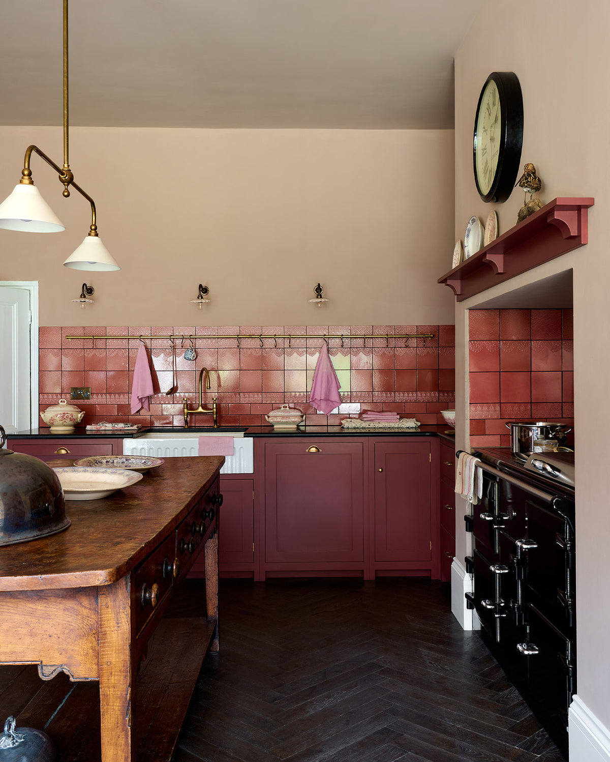 Looking into the sink run of a humble Refectory red shaker kitchen featuring glazed cupboards, a full-height pantry cupboard, a full-height cupboard and a large gas cooker in a chimney breast. The kitchen’s striking visuals are the Red Lace Market tiles used as a splashback in the chimney breast and along the sink run, enhancing the room's warmth. The space is decorated with vintage furniture, particularly an old, solid oak prep table.
