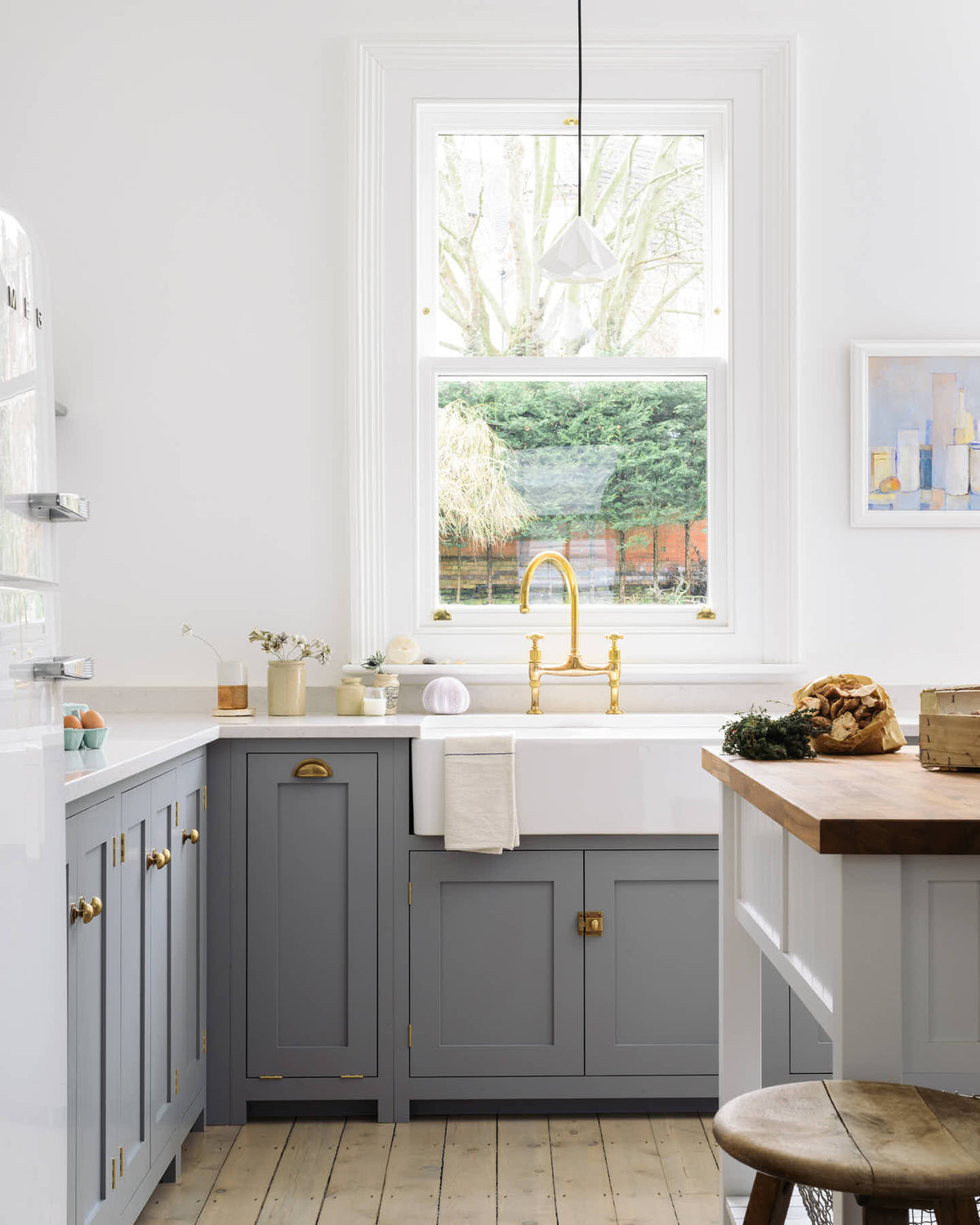 A light and bright Shaker kitchen features an L-shaped sink run with a double sink, an aged brass Ionian tap, silestone worktop, and a Danmask prep table with a solid oak worktop. Beautifully decorated with soft still-life paintings, a white refrigerator, a modern brass chandelier, and vintage furniture, it creates a wonderfully warm and contemporary space.