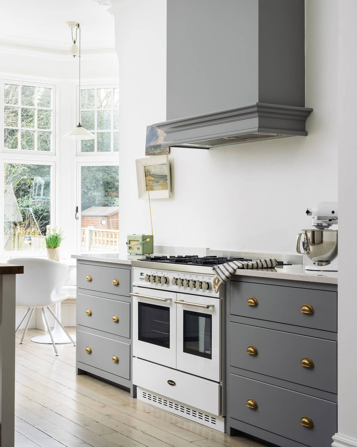 An oblique view of a light and bright Shaker kitchen features a cooker run with a canopy, Silestone worktop, and a Danmask prep table with a solid oak worktop. Beautifully decorated with soft still-life paintings, vintage furniture, and a breakfast nook, it creates a wonderfully warm and contemporary space.