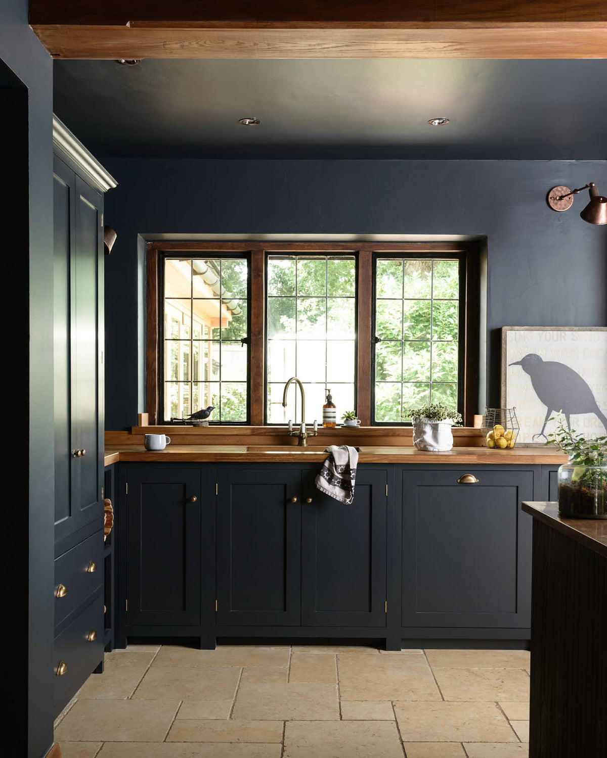 The Pantry Blue shaker kitchen features a sink run that is complemented by a vintage island with an Iroko wood countertop. Light stone flooring enhances the space, which includes wooden ceiling accents. The kitchen overlooks a large window that offers a stunning view of the surrounding trees and foliage.