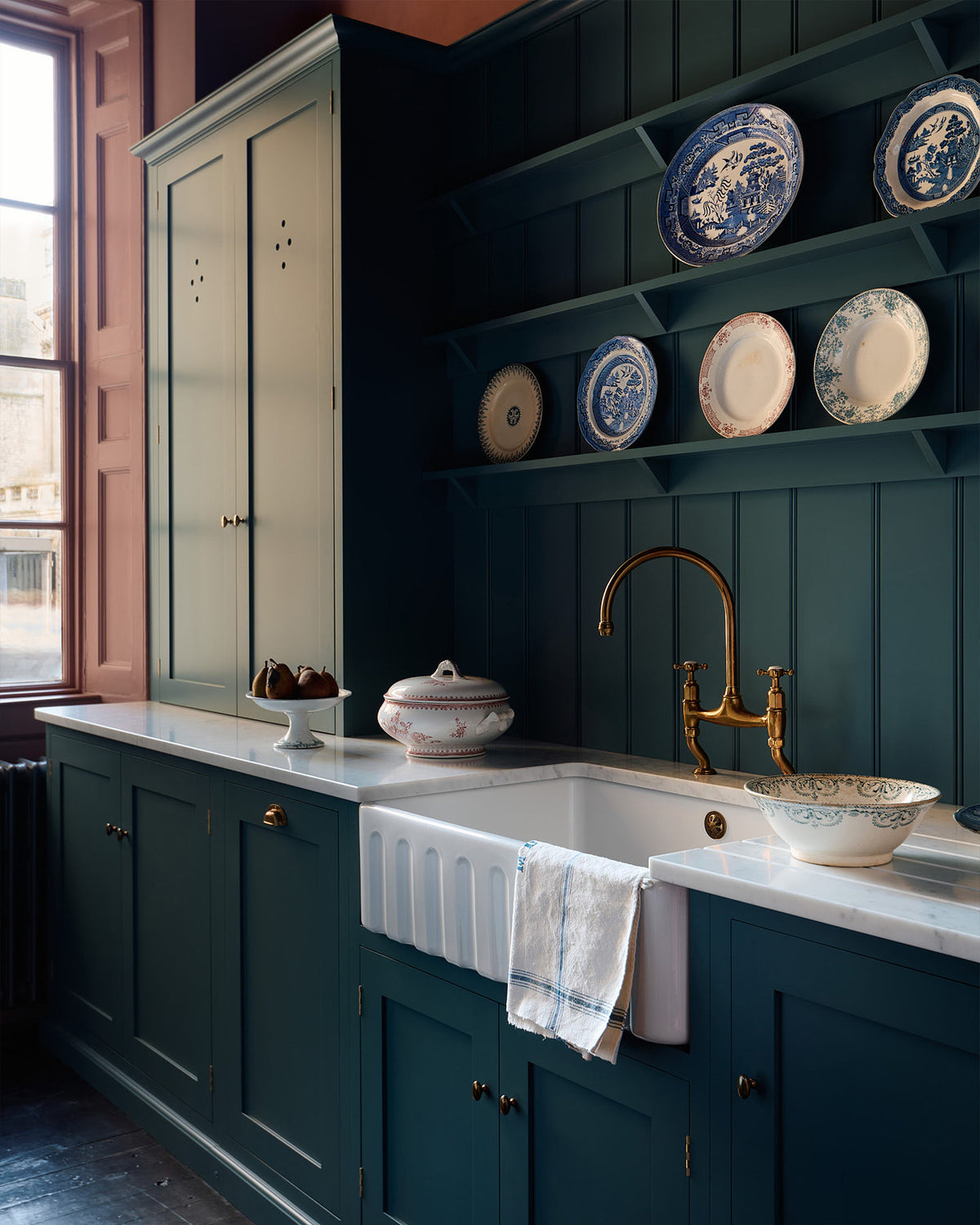 An oblique view of a sink run which shows the furniture, fittings and styling in more detail. The knobs are small, delicate and oval-shaped, and the integrated dishwasher to the left of the sink has a classic cup handle in the centre of the top rail. The space has an almost regal feeling with the moody wine walls, deep blue tones and the rich gold of the aged brass tap and fittings.