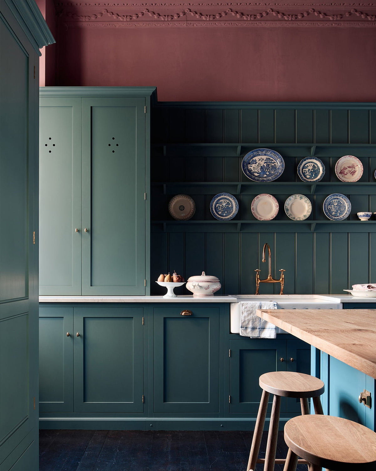 A shaker kitchen’s sink run with a countertop cupboard and panelled wall beside an original sash window. A small island with a rustic oak worktop stands at the centre, accompanied by a row of understated oak Helen’s Stools. Behind them, a grand freestanding pantry from our Classic English range is accented with Victorian-style brass Butler’s Knobs.