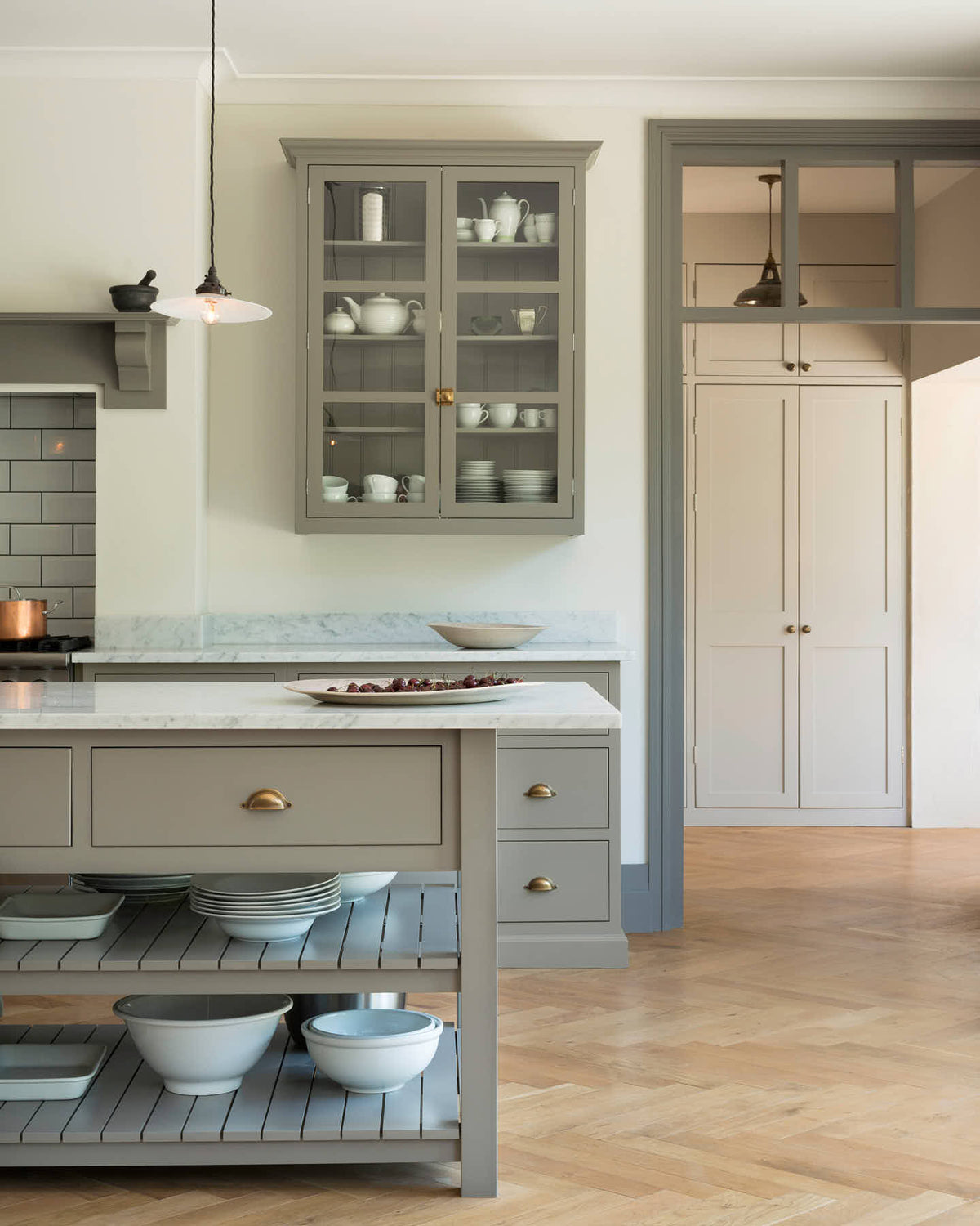 A grand warm-grey Shaker kitchen features a cooker run with a Carrara marble worktop, glazed wall cabinets, an overmantle canopy, and a wide kitchen island with a single sink. Adorned with beloved furniture, simple white platters, glass pendant lights, and pale wood herringbone floors, the space feels both sophisticated and cool.
