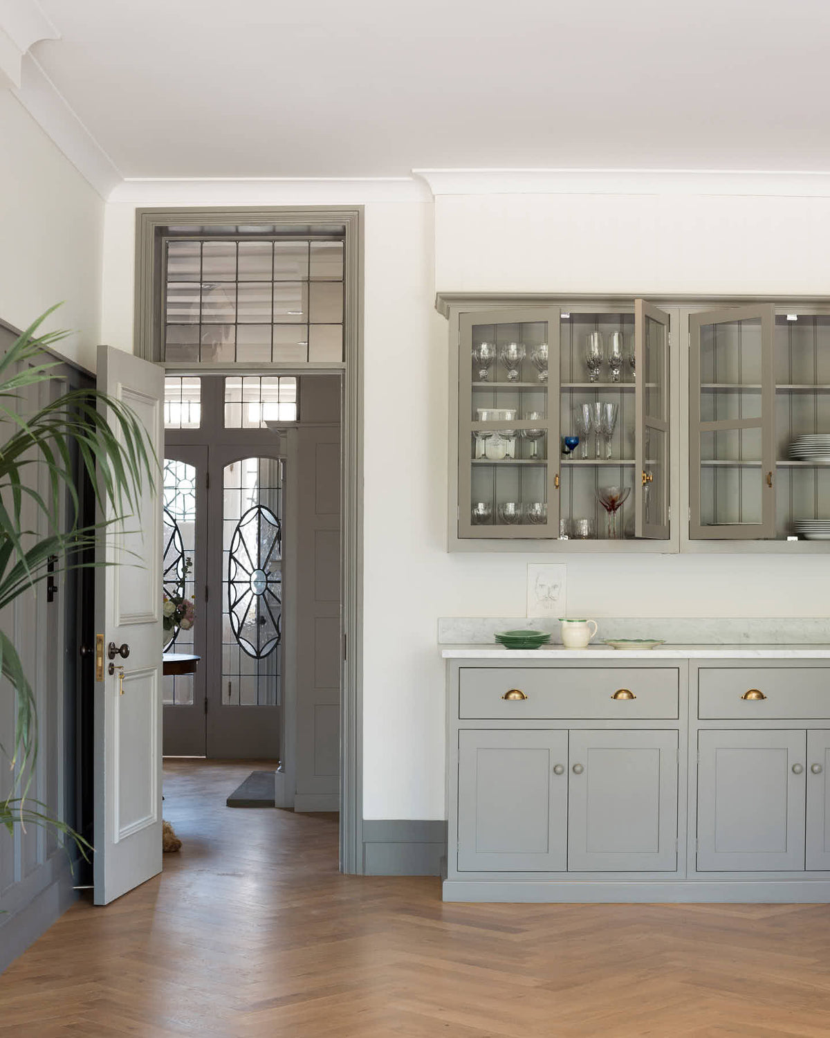 A quiet corner of a grand warm-grey Shaker kitchen features a pantry run with a Carrara marble worktop and glazed wall cabinets. Adorned with beloved furniture, simple white platters, and pale wood herringbone floors, the space feels effortlessly sophisticated and cool.