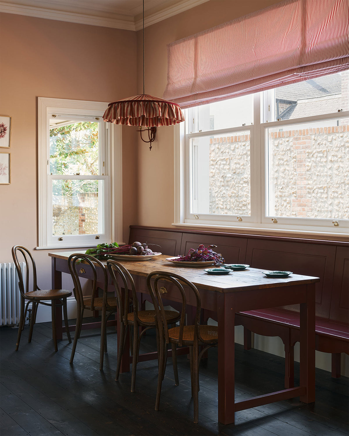 An angled view of a grand, refectory red dining room with a Millhouse refectory table, Bentwood café chairs and a bench. The table is adorned with red beets and leaves, sea-green heirloom platters, a vintage red-and-white striped chandelier, and red-rimmed wall lights.