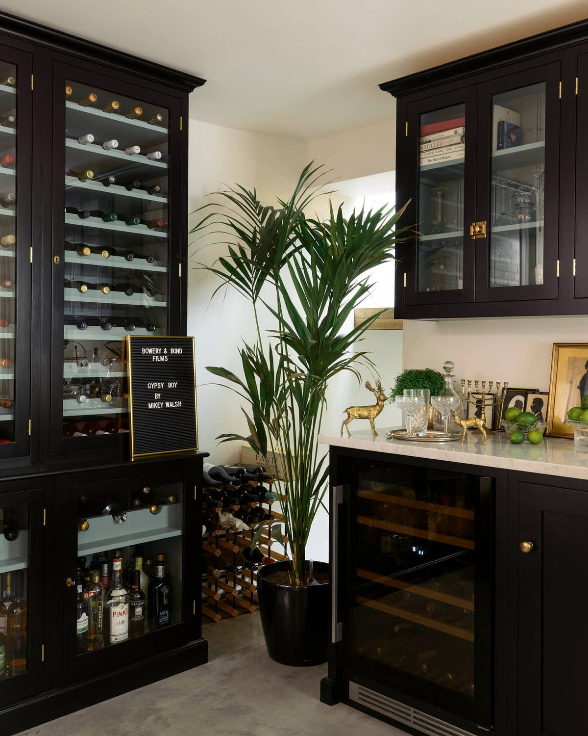 A classy cellar bar features a bank of curiosity cupboards, glazed wall cupboards, and a small wine cooler run with a Carrara marble worktop. Adorned with golden embellishments such as candelabras, a cake stand, deer ornaments, and a golden-framed gentleman's silhouette illustration, it creates a sophisticated and social space.