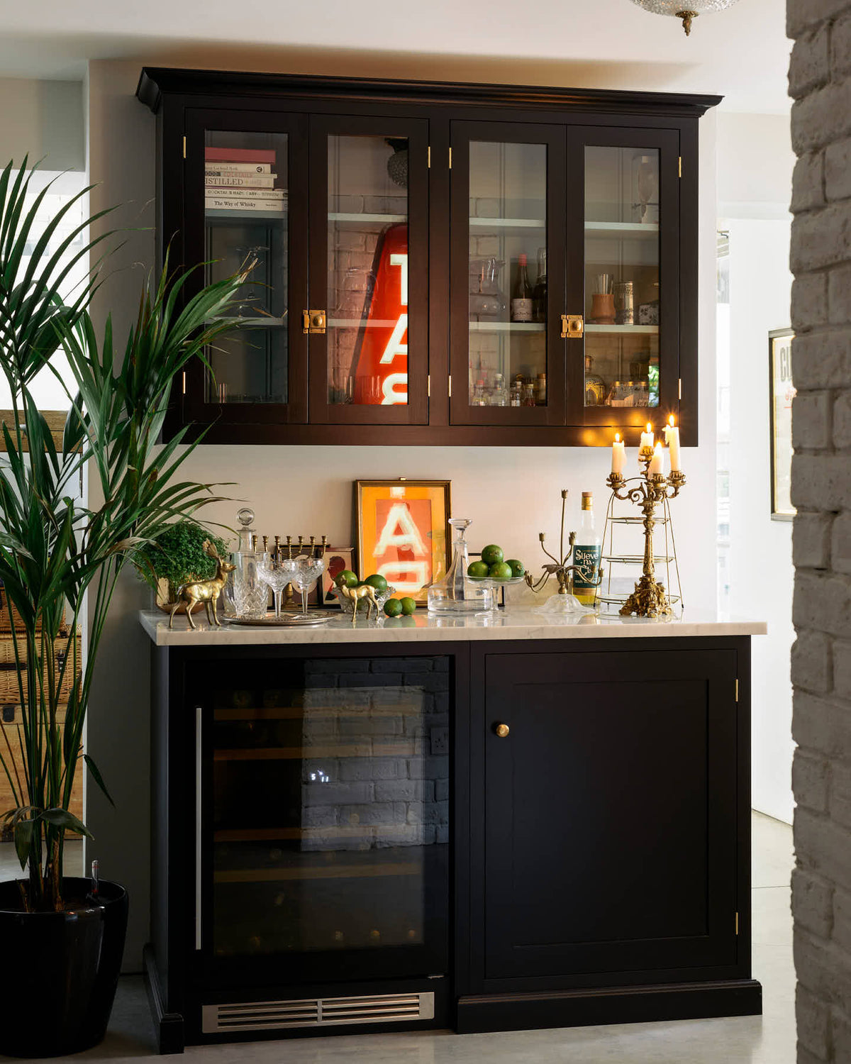 A classy cellar bar features glazed wall cupboards and a small wine cooler run with a Carrara marble worktop. Adorned with golden embellishments such as candelabras, a cake stand, deer ornaments, and a vintage red Tabac neon lights, it creates a sophisticated and social space.
