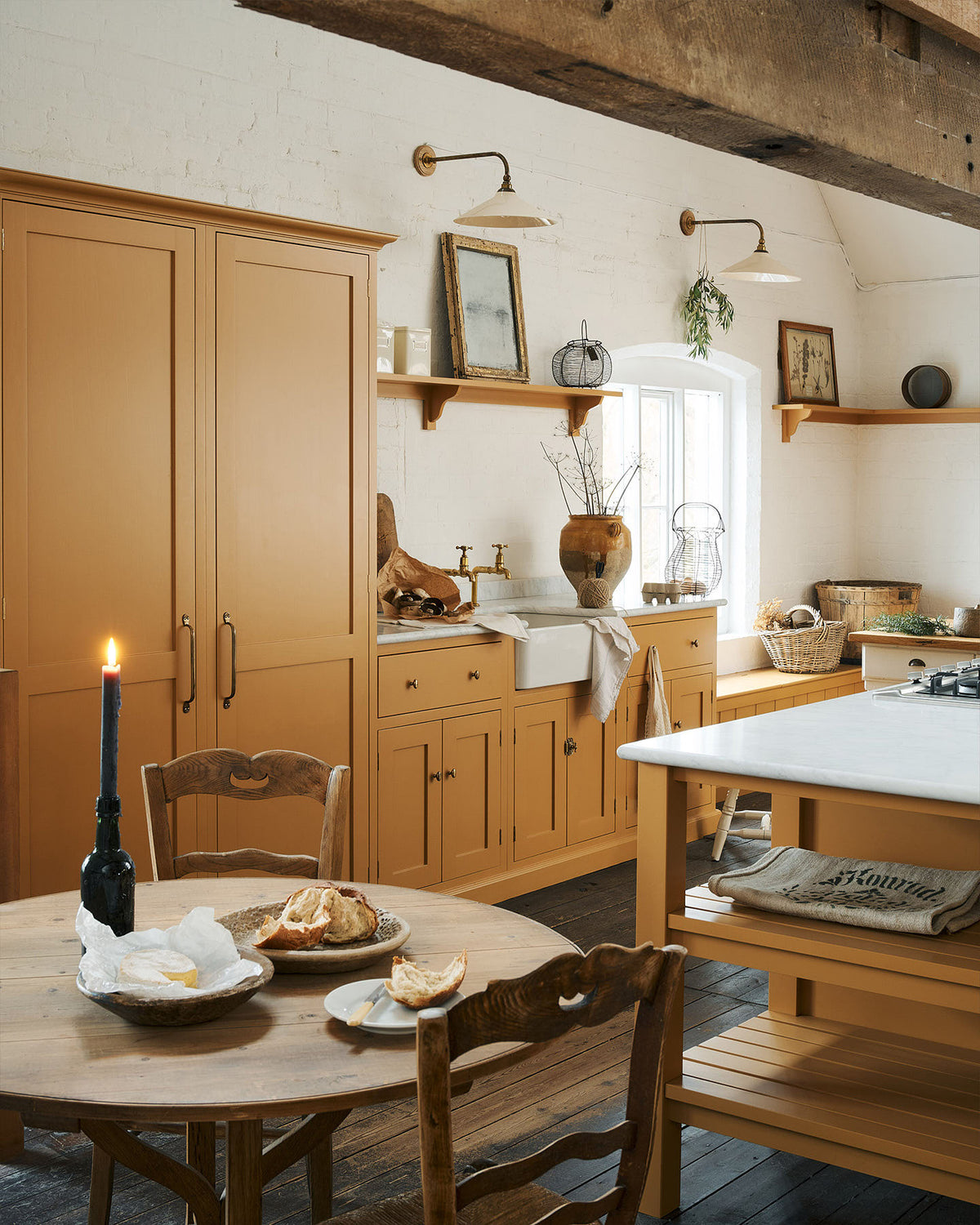 An oblique view of a scullery yellow shaker kitchen with a small sink run with a Carrara marble sink, a full-height cabinet, shaker shelves, an L-shaped shaker bench seat and a kitchen island. The kitchen features a single Belfast sink with aged brass Mayan taps, a large vineyard table, a collection of vintage furniture and beloved items such as mixing bowls and fresh produce. The ceilings are high and retain their original exposed wooden structure.