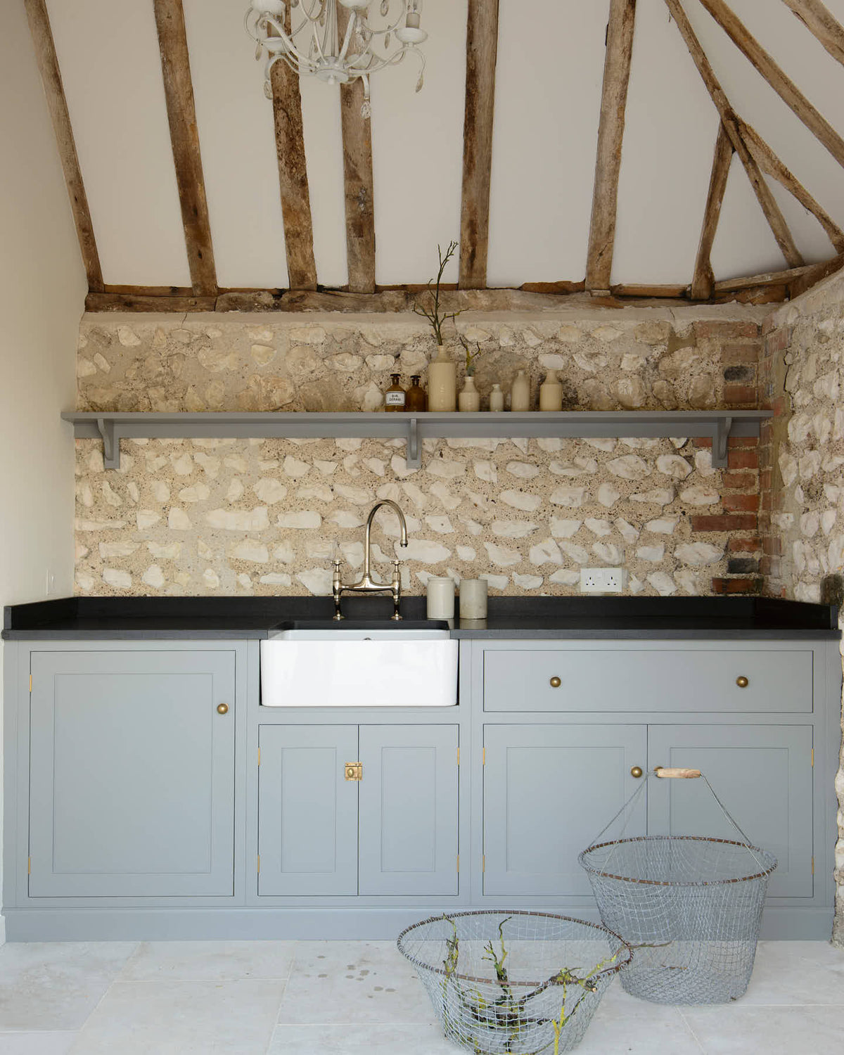 A chic and simple muted-blue sink run against the original wall textures of the pool house features a single Belfast sink with a nickel Ionian tap, a black honed granite worktop, and a long shelf. Adorned with vintage ceramics and brass fittings, it creates a functional corner in the space.