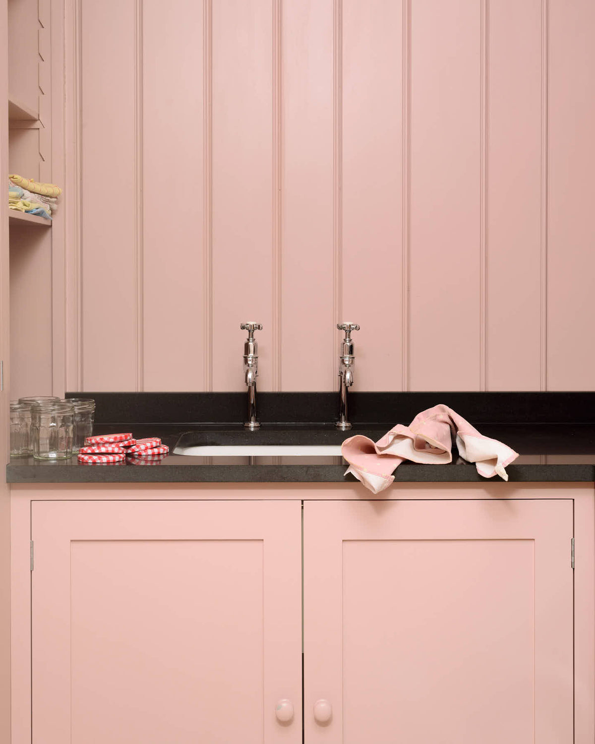 A chic and simple muted-pink laundry room features a sink run with a honed black granite worktop, a full-height cupboard, a single sink, and nickel Mayan taps. Adorned with empty jam jars, folded tea towels, and a black washing
machine, it creates a functional corner in the space.
