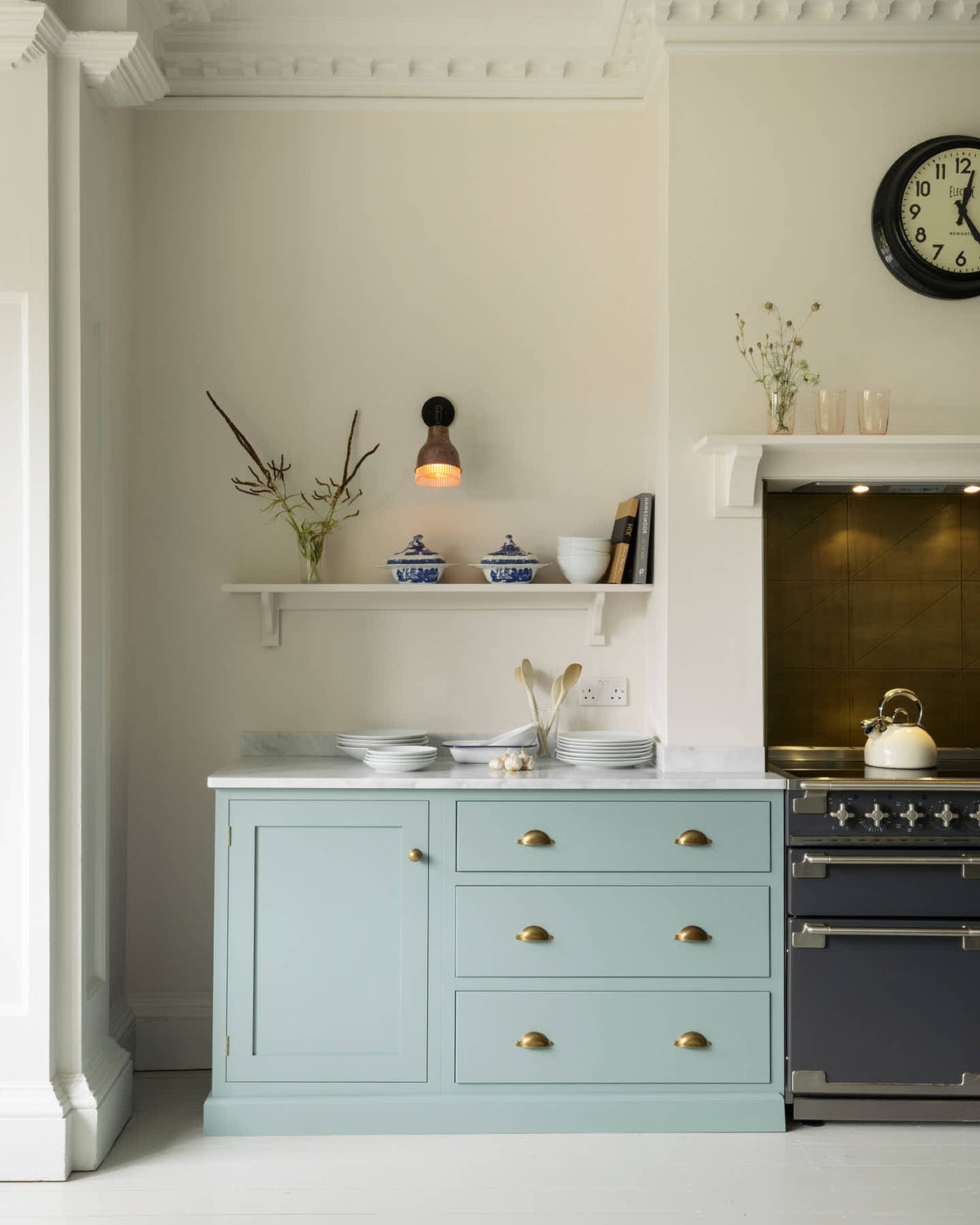 In a quiet corner of a minimal Trinity Blue shaker kitchen, the cooker run features a marble worktop. There are assorted ceramics, porcelain dishes, and books placed on the worktop and shelves.