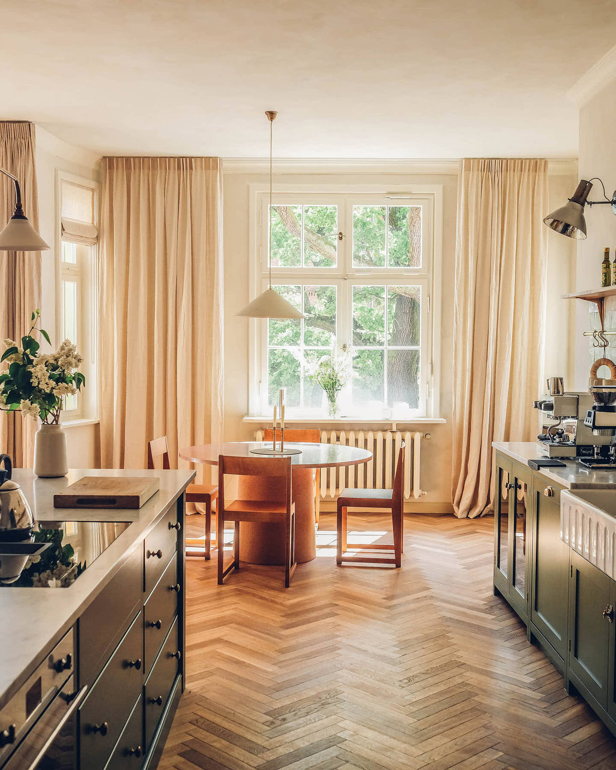 A warm, glowy image of a mid-century style dining area at the bottom of a traditional Shaker kitchen. A large window at the back of the room creates a pool of beautiful, summery light whilst wooden parquet floors, limewash walls and floor-to-ceiling curtains add layers of texture.