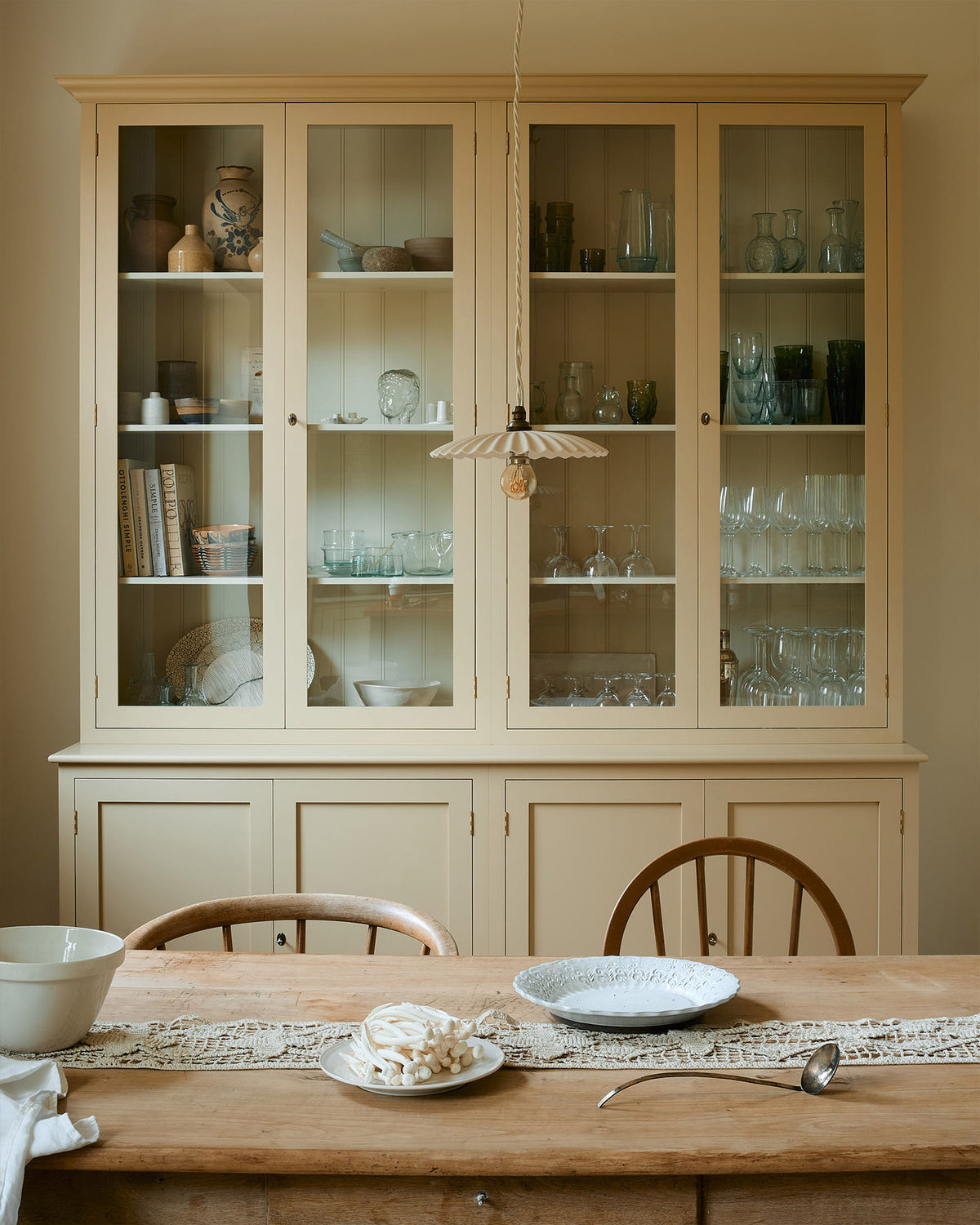 A cream, glazed pantry cupboard stands proudly at the back of a shaker kitchen. In the foreground is an old pine table and antique stick-back chairs, styled with vintage lace, an antique silver spoon and deVOL’s heirloom round platter. A delicate frilly pendant light hangs above the table.