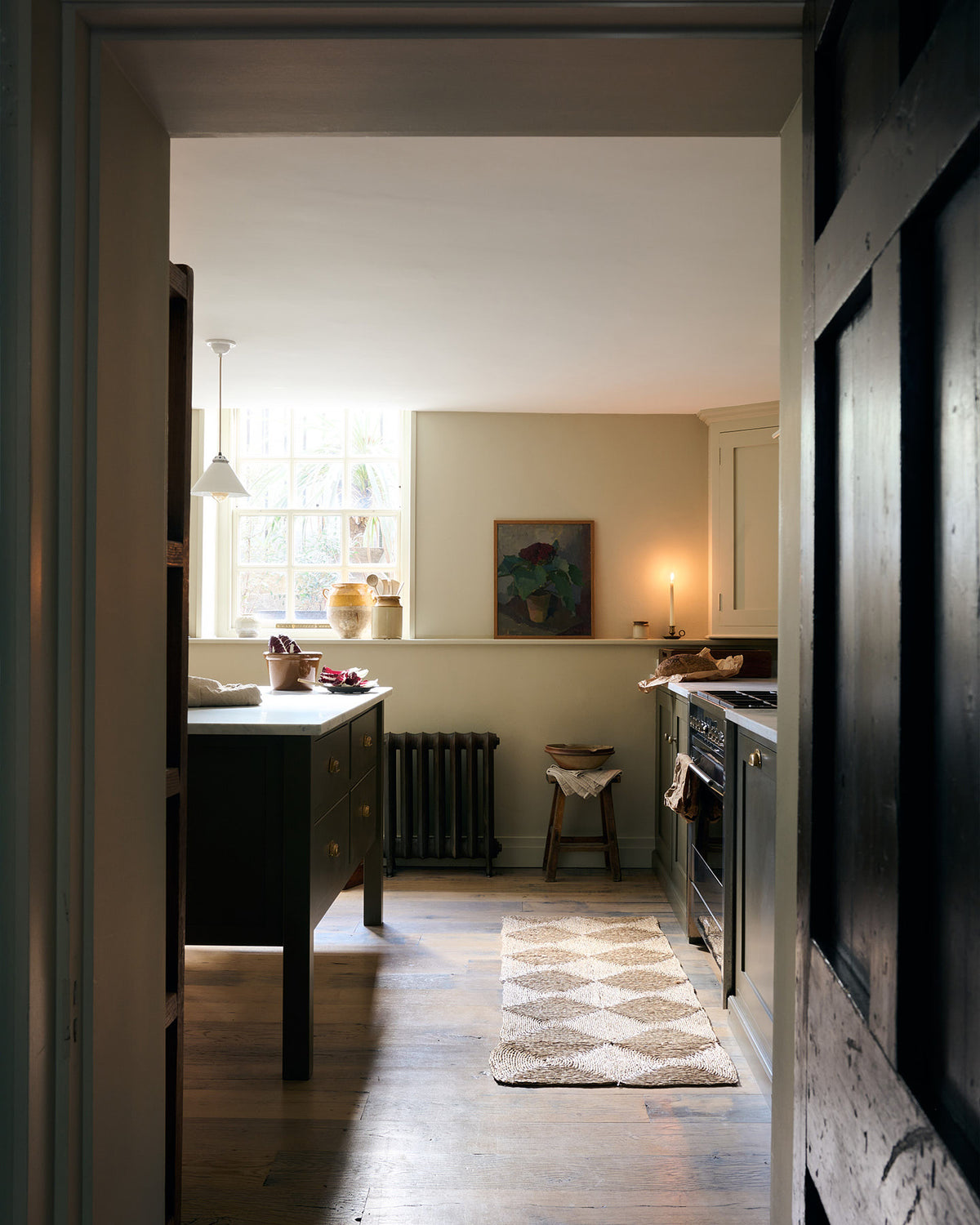 Looking through a dark corridor, a well-lit olive green Shaker kitchen comes into view, featuring a sink and cooker run, along with a freestanding prep table topped with Carrara marble.