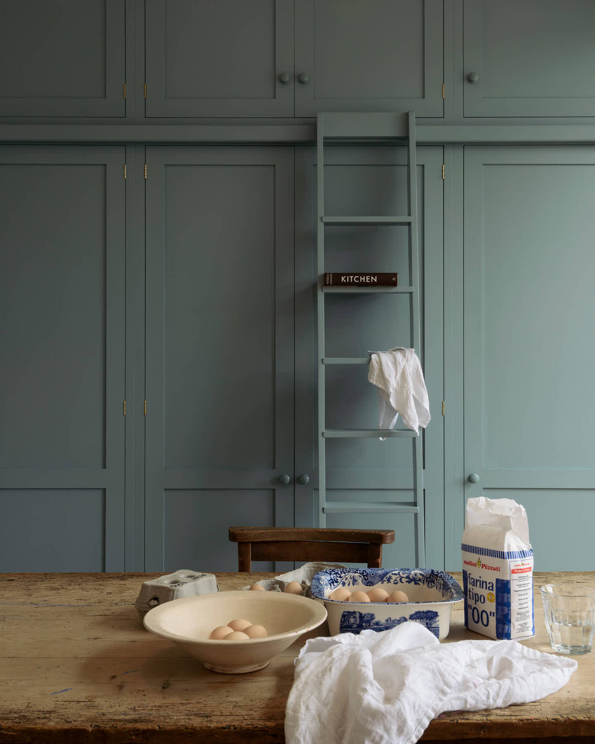 A floor-to-ceiling run of cabinetry, a library ladder propped up to reach the highest cupboards and simple wooden knobs, all painted in a soft, duck egg blue. In the foreground is a wooden table with a wonderfully worn and knotted top. Flour, egg, and some linen napkins set the scene for an evening of baking.