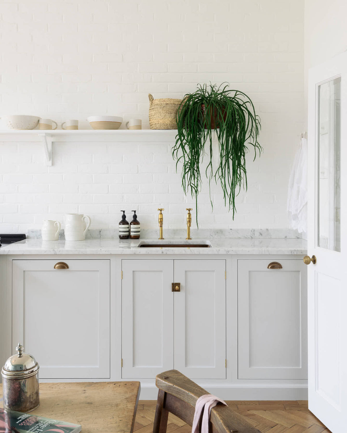 A closer-look at a beautiful Trinity Blue Shaker kitchen featuring a cooker and sink run, a vintage wooden dining table with a wonderfully worn top, and a single copper sink with aged brass Mayan taps. A long Shaker shelf spans above the kitchen area, complemented by a low, hanging house plant, antique furniture, and grand solid oak herringbone floors.