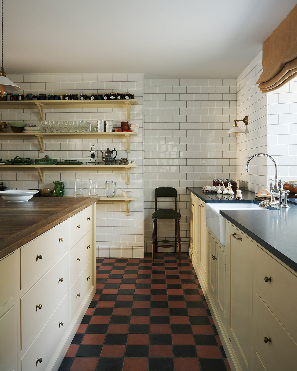 A moody and atmospheric butter-yellow painted shaker kitchen with a simple, utilitarian feel. Light cupboards and glossy, white tiled walls contrast with original black and red chequerboard quarry tiles. The scullery features a central island, a long run of base cabinets that includes a double sink unit, honed back granite worktops and fitted Shaker shelving.