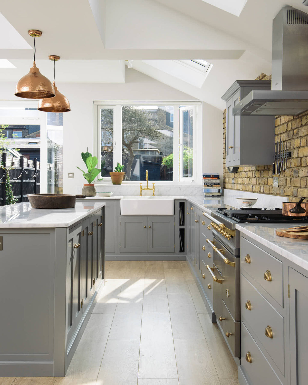 Nestled between a soft industrial grey L-shaped sink and cooker run, which features a Carrara marble worktop, wall cupboards, and a kitchen island, is a single Belfast sink with an aged brass Ionian tap. Adorned with a weathered brick wall installation, copper pendant lights, simple ceramics, and colourful books, it creates a modern and functional kitchen for enthusiastic food connoisseurs.