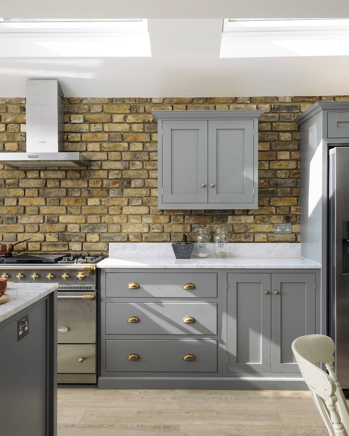 A soft industrial grey Shaker kitchen features an L-shaped sink and cooker run with a Carrara marble worktop, a kitchen island, wall cupboards, and a large silver refrigerator. Adorned with a steel canopy, a weathered brick wall installation, a cream gas cooker, and a stone pestle and mortar, it creates a modern and functional kitchen for enthusiastic food connoisseurs.