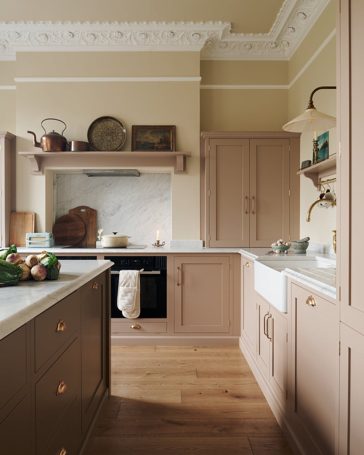An elegant and soft Princelet pink Shaker kitchen unfolds with an L-shaped cooker and sink run topped in Carrara marble. Alongside countertop cupboards, a kitchen island with a breakfast nook, and an over mantle above the alcove of a chimney breast and marble splash back, it create a beautifully functional space.