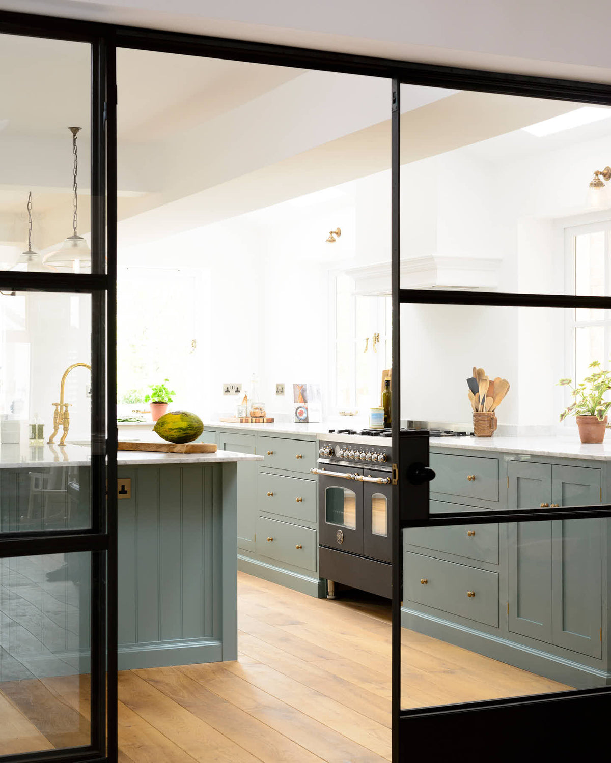 Past a black-framed internal glass sliding door is a bright and elegant Trinity blue Shaker kitchen featuring a long cooker run with a canopy, a kitchen island with a single sink, an aged brass Ionian tap, and a Carrara marble worktop. Beautifully decorated with artisan ceramics, books, wooden chopping boards, and a watermelon, it creates a humble family space.
