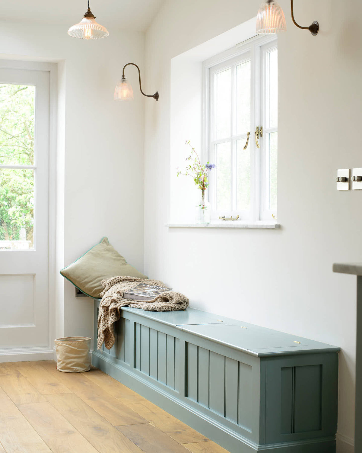A bright and elegant hallway to the garden features a vintage dresser and a long Shaker bench seat with storage compartments. Beautifully decorated with a vintage dresser, a glass-frilled pendant light, a swan-neck glass-frilled wall light, a pillow, a crocheted blanket, and a vibrant house plant, it creates a humble family space.