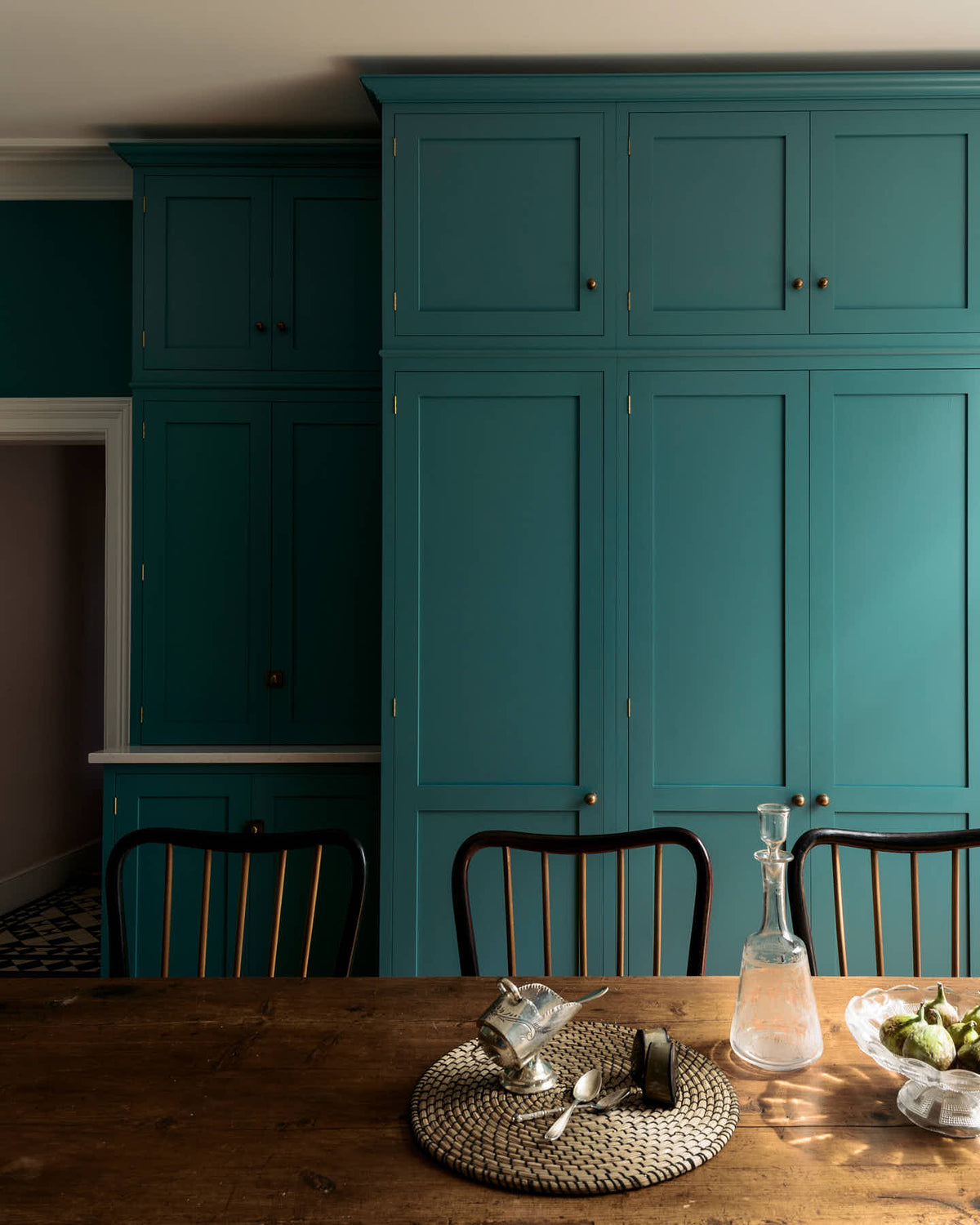 A grand, seafoam green Shaker kitchen featuring a bank of full-height pantry cupboards and a free-standing closed larder with Carrara marble worktop. Adorned with beautiful vintage furniture, a glass bowl of green figs, antique silverware, and a decanter, the kitchen reflects the refined tastes of its socialite and collector owners.