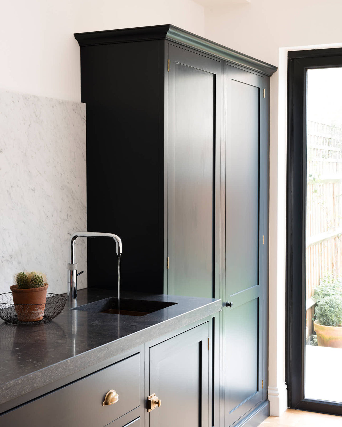 An oblique view of a bright and sophisticated pantry-blue Shaker kitchen features a sink and cooker run with a Belgian blue limestone worktop, a Carrara marble splashback and full-height cupboards. Adorned with a silver tap, a single copper sink, a cactus plant, and beautiful brass fittings, it creates a functional and contemporary kitchen space.