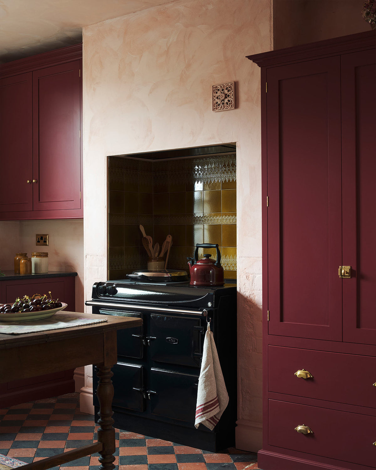 An oblique view of a warm and simply gorgeous refectory-red Shaker kitchen features a sink run with a black granite worktop, a wall cupboard, a pantry cupboard, and a gas stove in a chimney breast with Sepia Lace market splashback tiles. Beautifully decorated with vintage furniture, artisan ceramics, and a matching red teapot, it creates a humble and endearing space.