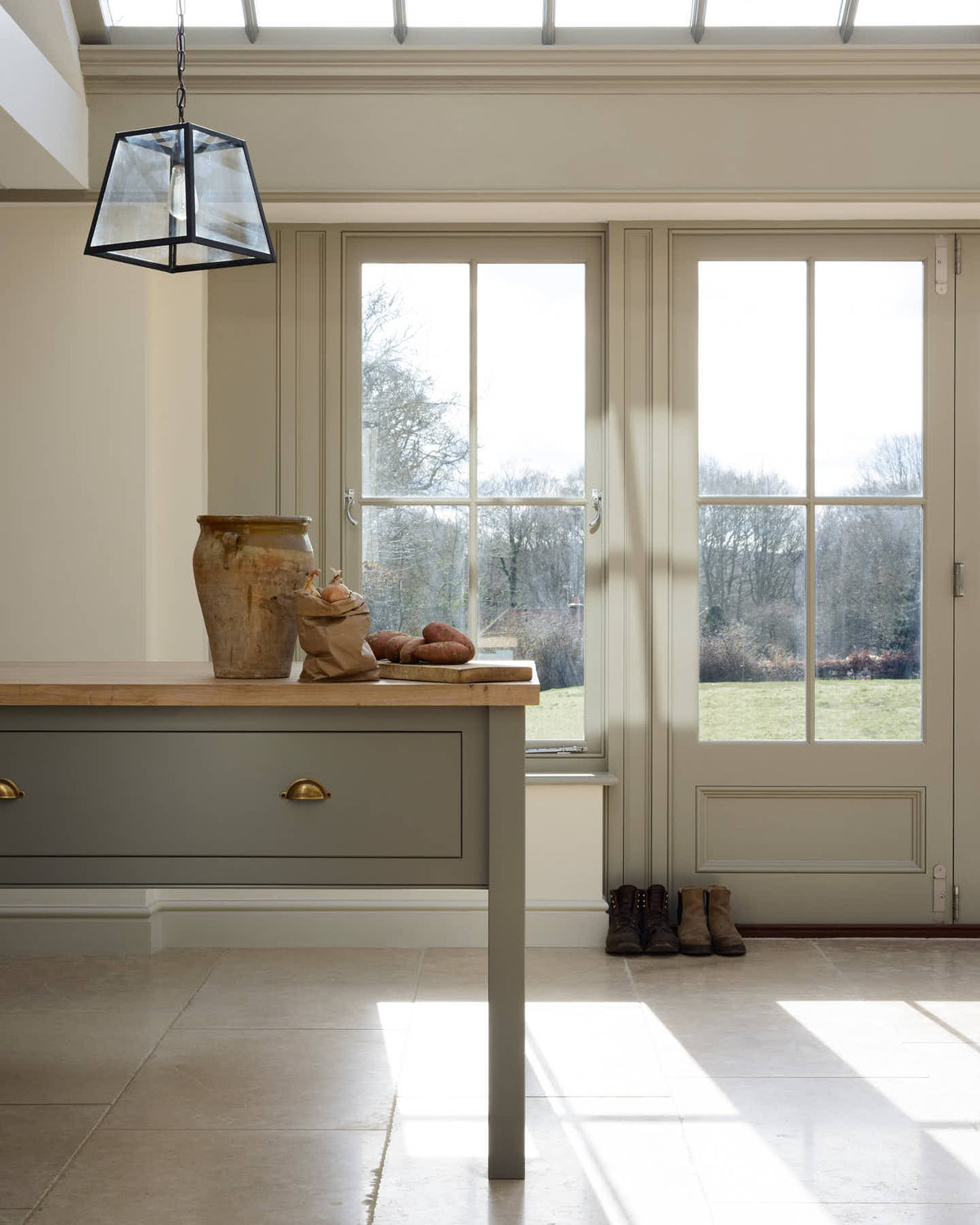 A light and beautifully calm mushroom-coloured Shaker kitchen, under a skylight and bifold patio doors, features a muted blue bespoke prep table with a solid oak worktop. Adorned with steel-framed hanging lights, vintage ceramics, sweet potatoes, a brown bag of onions, and muted, textured natural stone, it creates a lovely minimalist kitchen and living space.