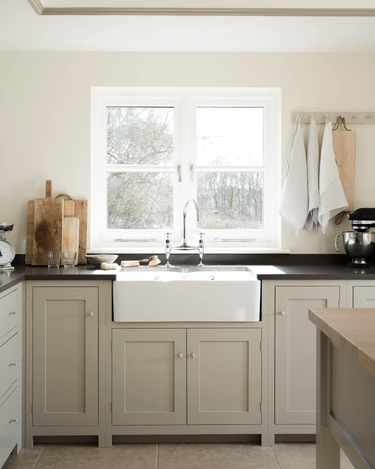 A closer look at a light and beautifully calm mushroom-coloured Shaker kitchen’s L-shaped sink and cooker run with a black granite worktop, a double sink with an antique silver Ionian tap. Adorned with wooden chopping boards, a silver scale, a black mixer, tea towels, and muted, textured natural stone, it creates a lovely minimalist kitchen and living space.