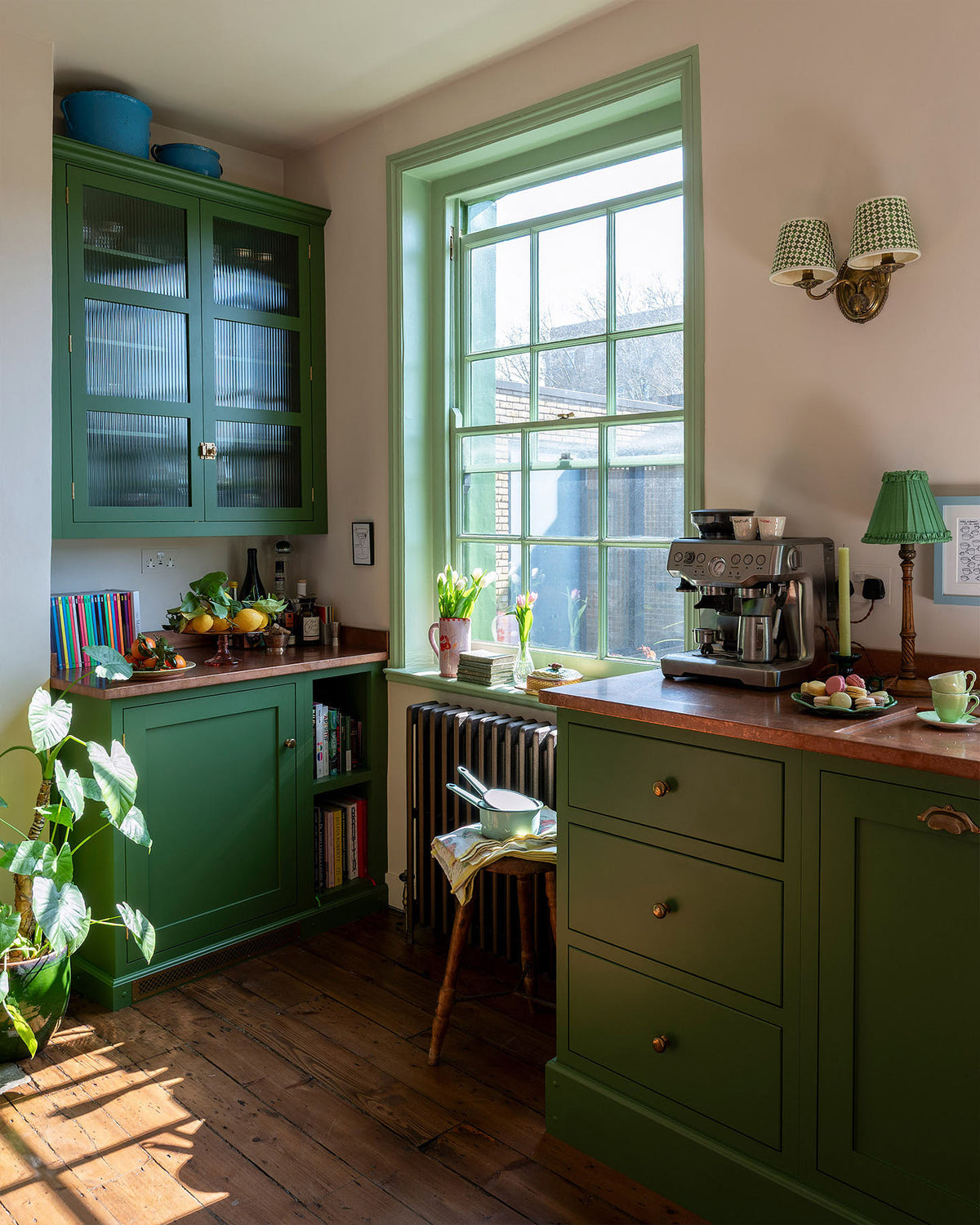 A corner of a bright and humble lime-green kitchen with an L-shaped cooker and sink run topped with aged copper worktops, a glazed counter cabinet, and colourful accents like tulips, a bowl of lemons, macaroons, books and assorted bottles of alcohol.