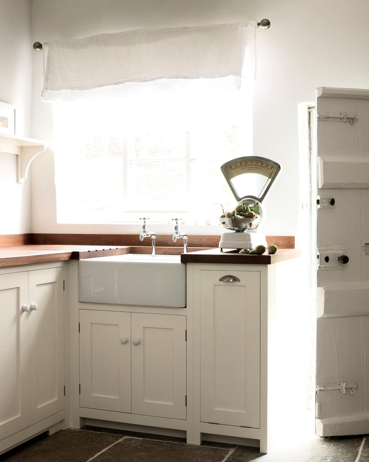 Minimalist white linen Shaker kitchen with a dark sapele wood worktop, Belfast sink, and antique silver taps. Bright light pours in from the garden door, highlighting vintage scales with green pears.