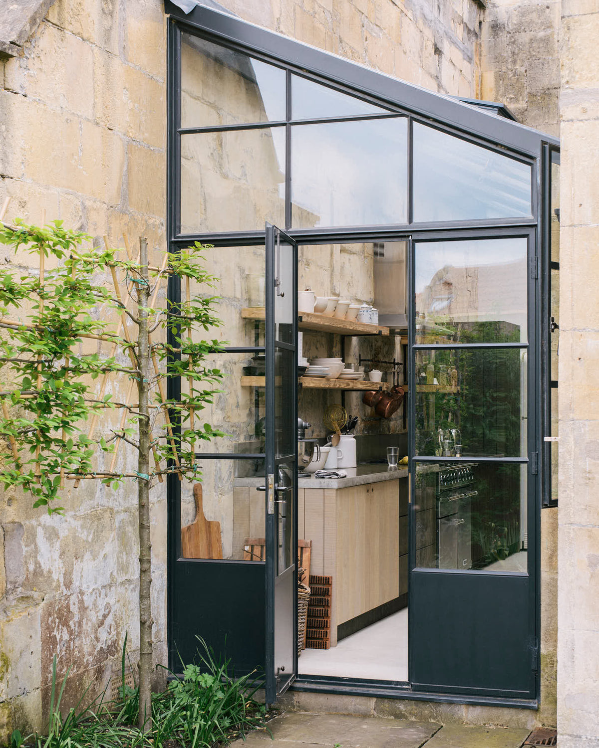 A quaint up-and-over glass design sits between a historic Georgian house, revealing a fresh and calming Sebastian Cox kitchen adorned with a bespoke moss green colour tint, copper pans, and artisan ceramics. This space creates a practical earth-toned environment that complements the original limestone walls.