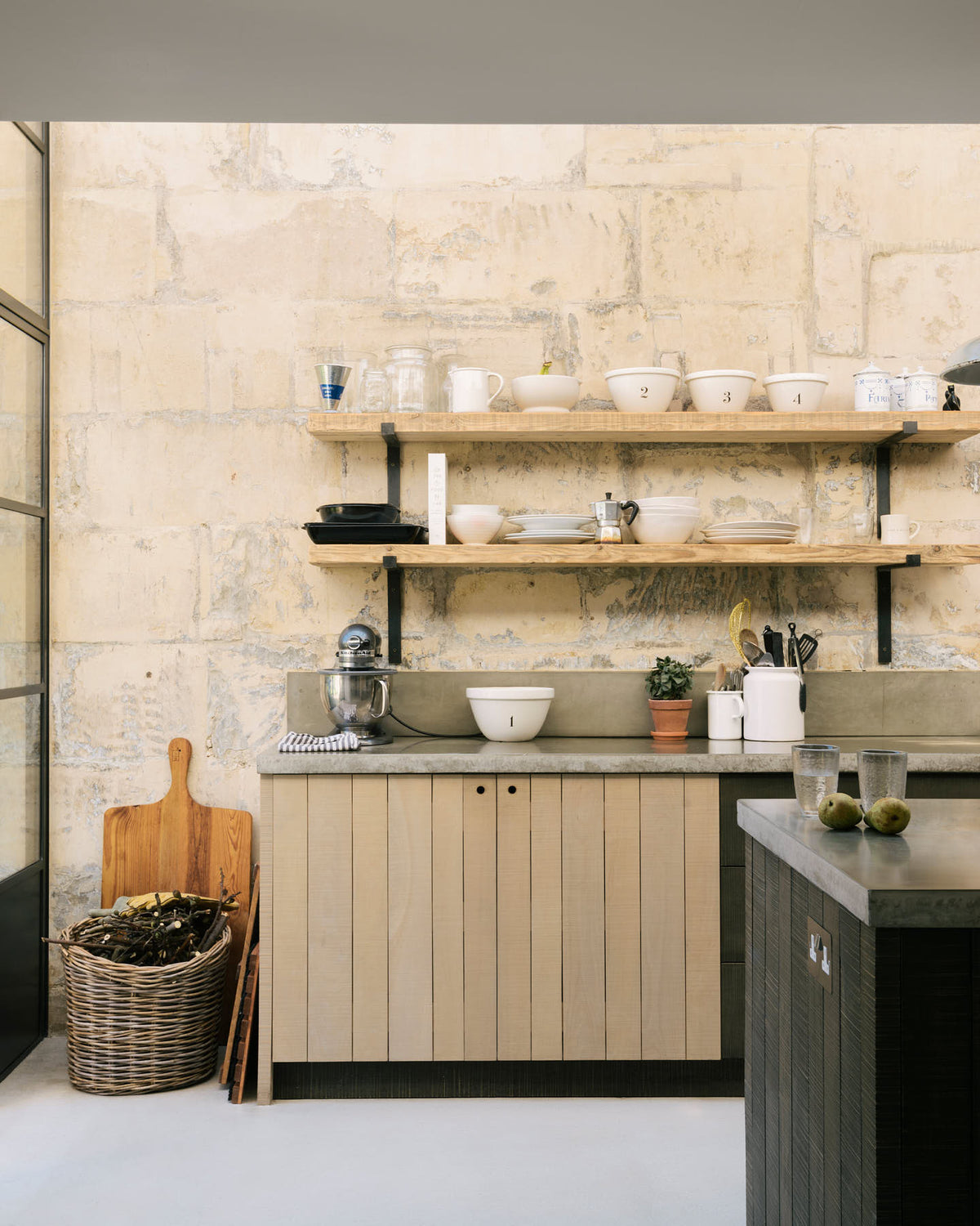 A quiet corner of a fresh and calming Sebastian Cox kitchen features a long cooker run with a cement worktop and splashback, and a kitchen island. Adorned with a bespoke moss green colour tint, large chartreuse board, artisan ceramics, a wicker basket, and a stainless steel mixer, this kitchen creates a practical earth-toned space that complements the original limestone walls.